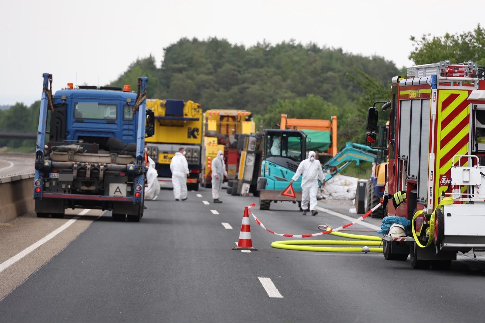 Feuerwehrleute in Schutzanzügen laufen über die Autobahn. Im Hintergrund ist der umgestürzte Anhänger zu sehen.