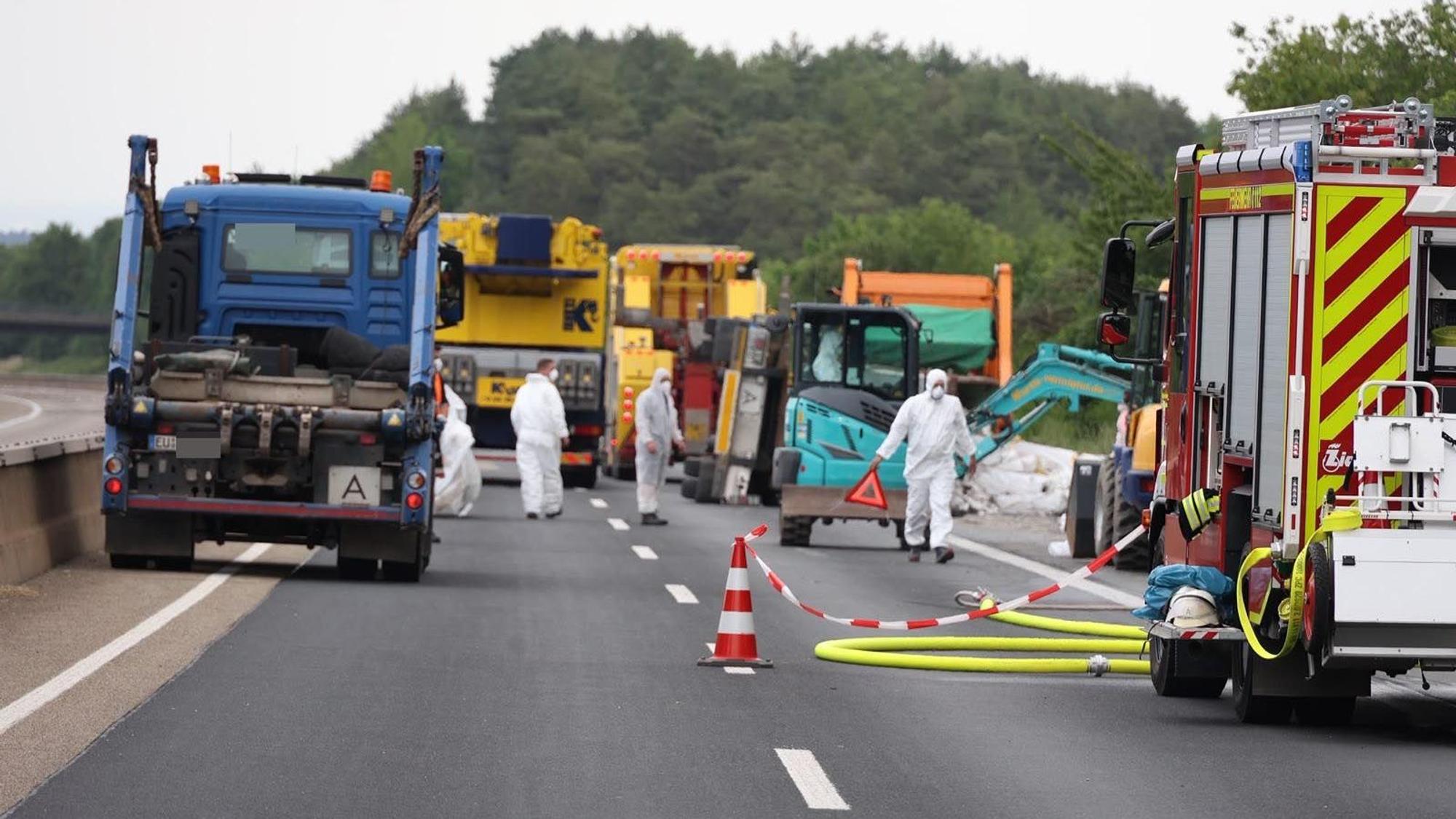 Einsatzkräfte in Schutzanzügen laufen über die Autobahn. Im Hintergrund ist der umgestürzte Anhänger zu sehen.