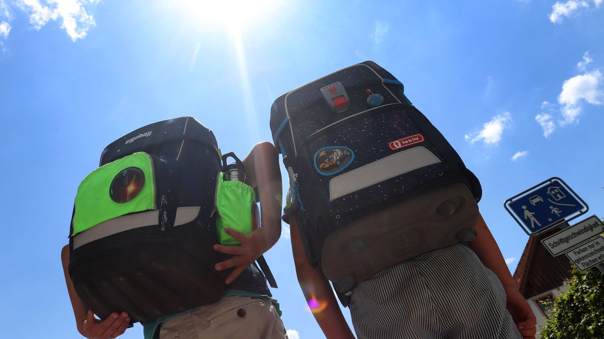 Zwei Kinder haben Hitzefrei und laufen mit ihren Büchertaschen im Sonnenschein nach Hause.
