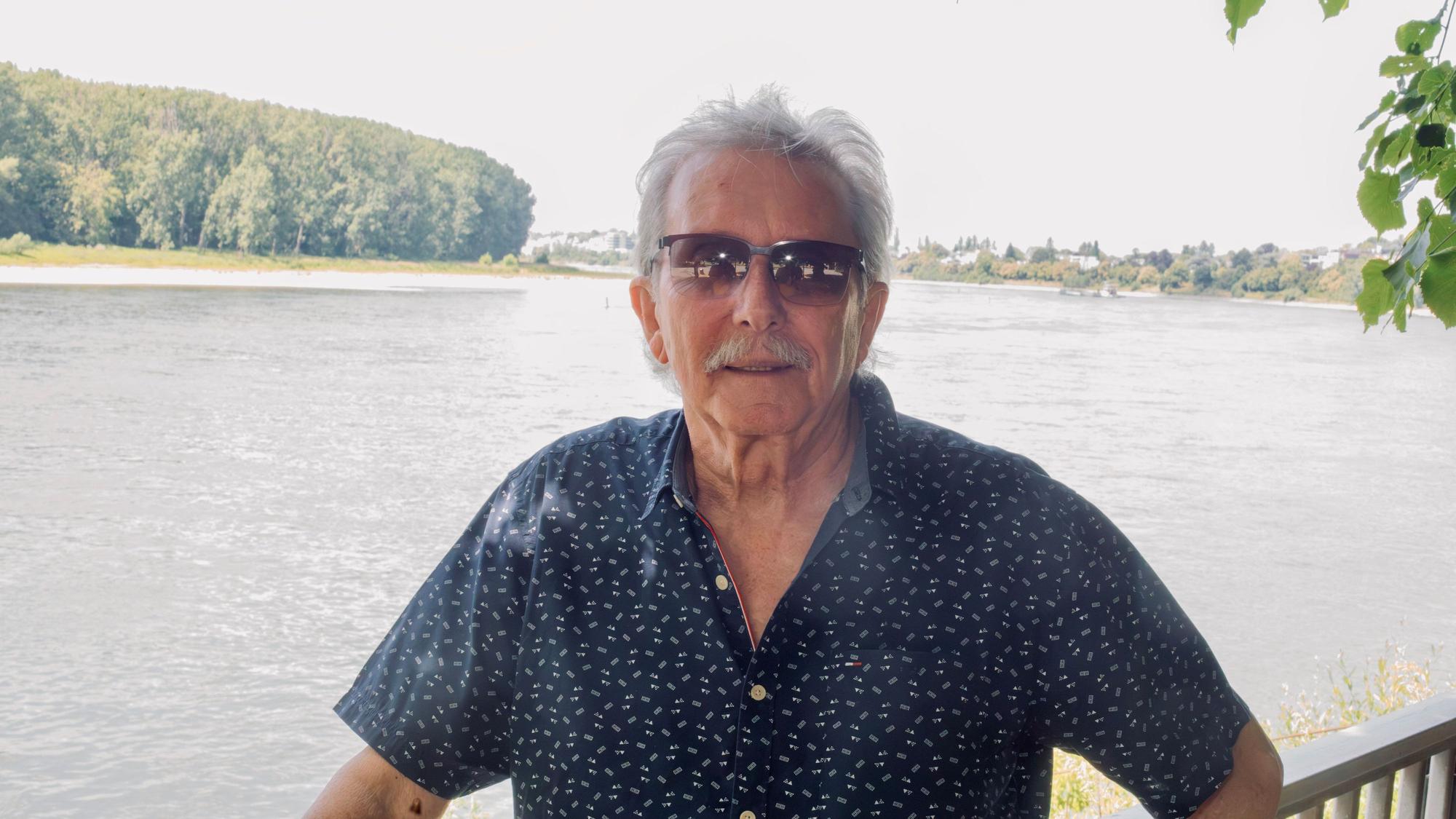 Ein Mann mit kurzem grauen Haar und Sonnenbrille und Schnurrbart steht vor dem Rhein in Porz.