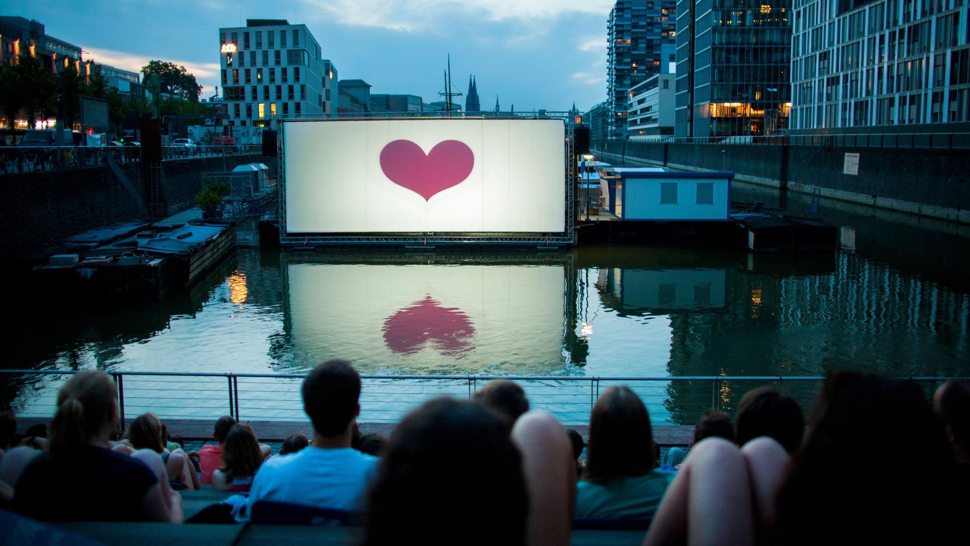 Im Open-Air-Kino am Rheinauhafen vor der Domkulisse warten Besucher auf die Filmvorführung. (Archivbild)