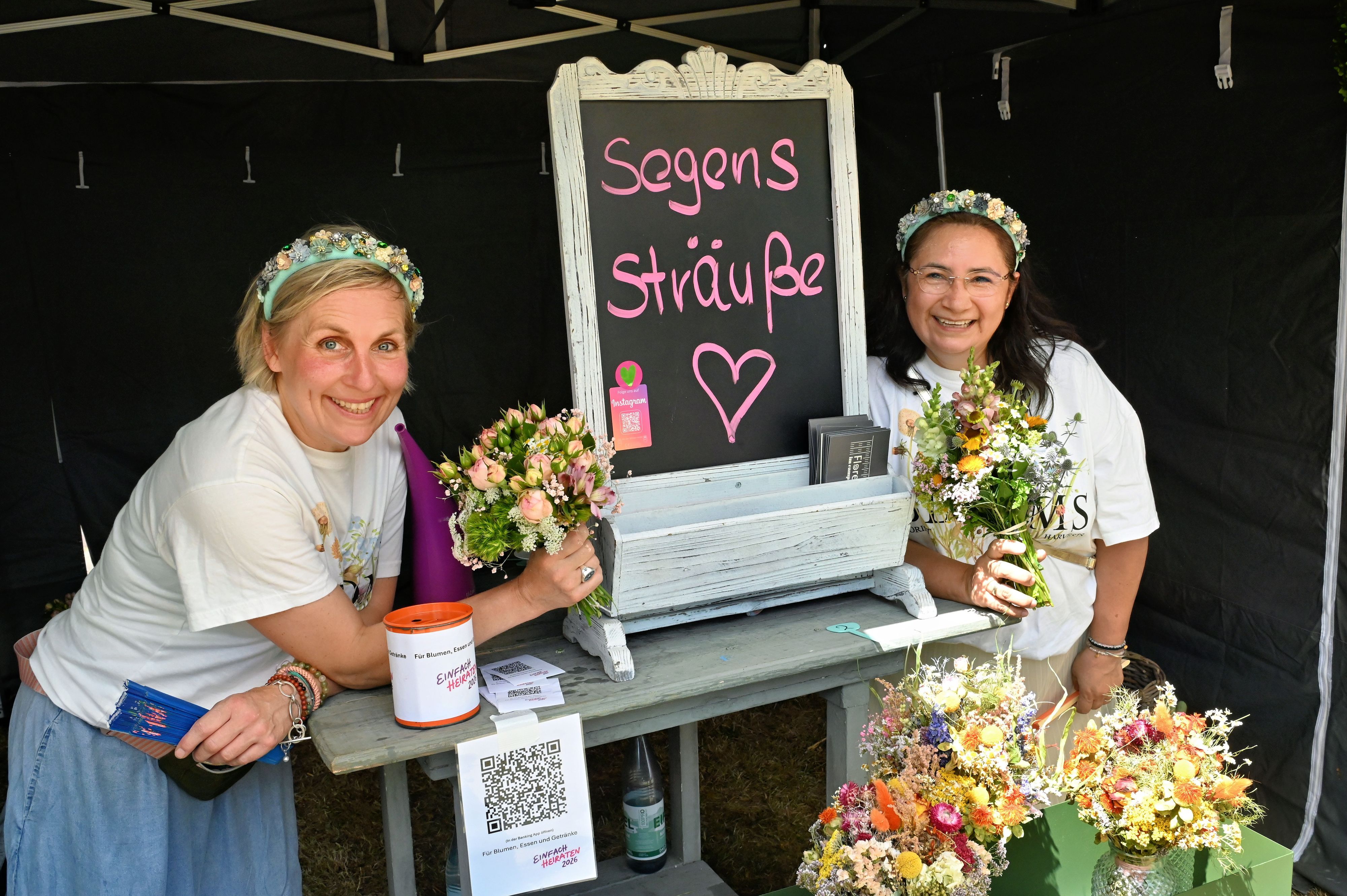 Die beiden Frauen mit Blumenkränzen im Haar halten Blumensträuße in die Kamera.