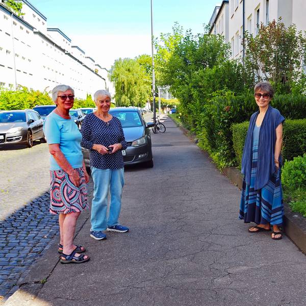 Drei Frauen stehen auf einem Bürgersteig. Ein Auto parkt hinter ihnen.