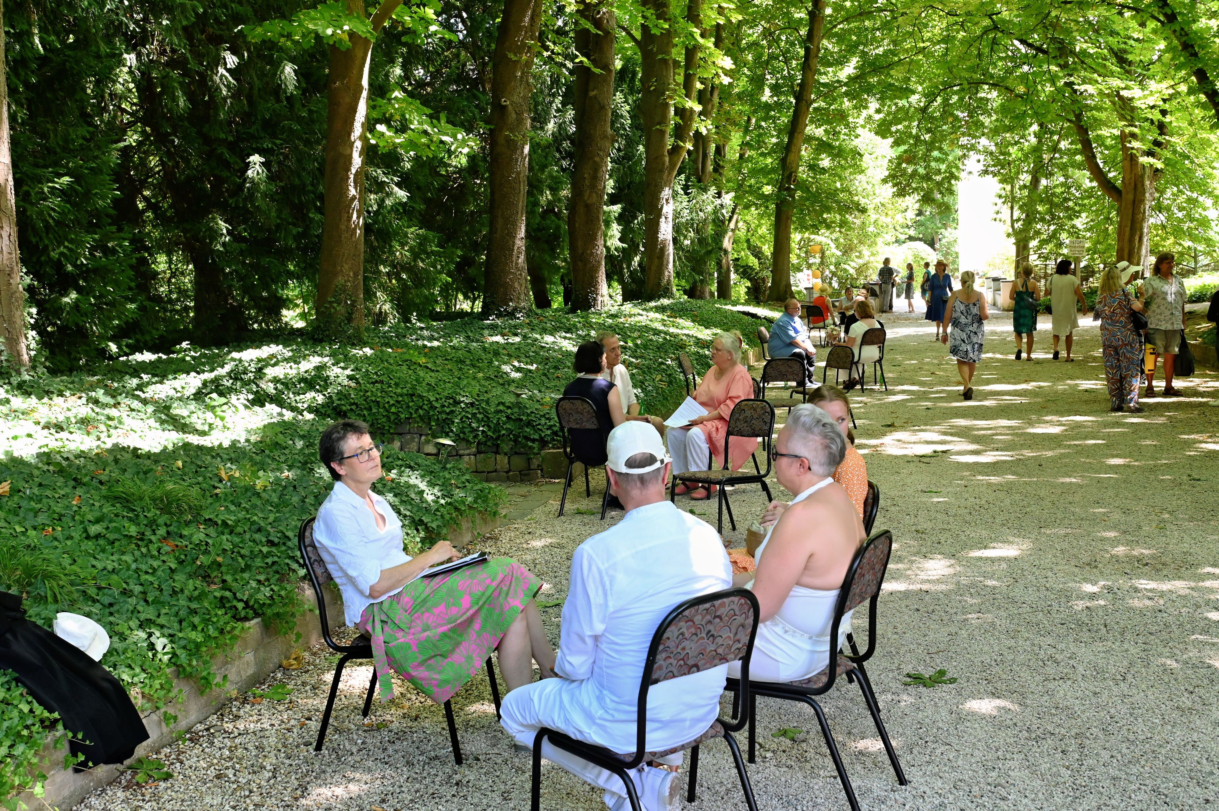 Menschen sitzen auf Stühlen auf einem Kiesweg unter Bäumen.