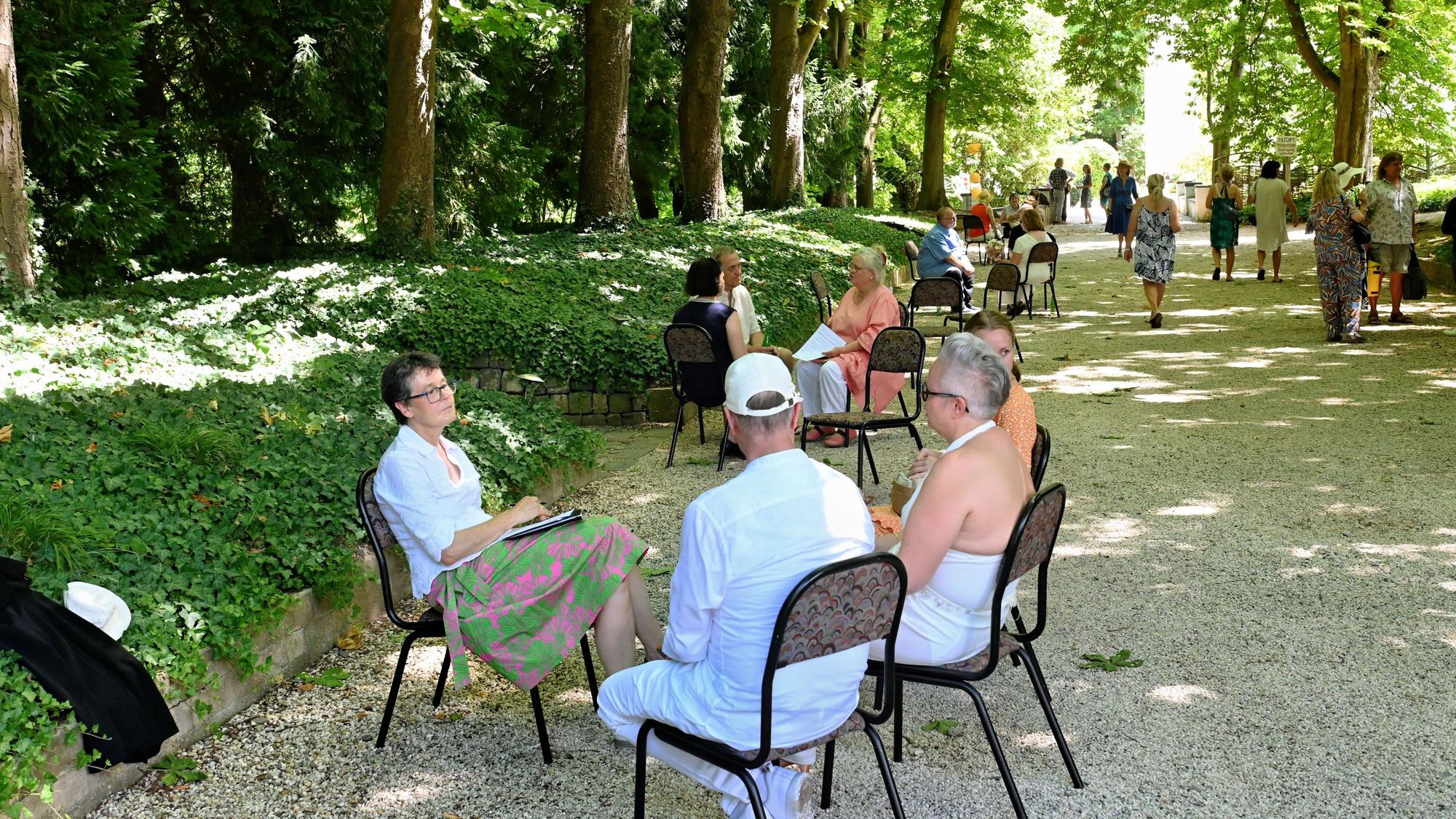 Menschen sitzen auf Stühlen auf einem Kiesweg unter Bäumen.