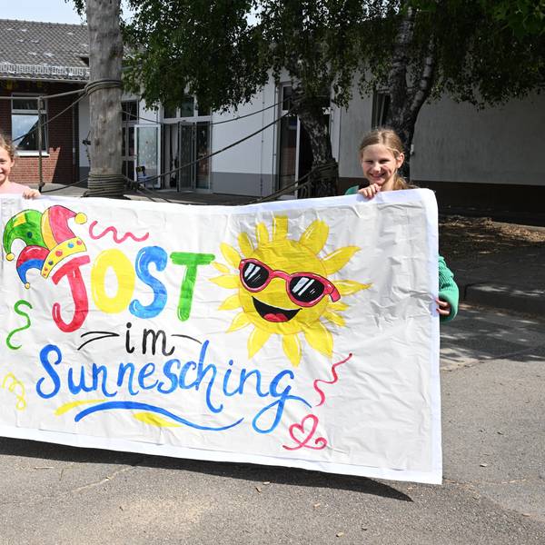 Zwei Grundschülerinnen halten ein buntes Plakat hoch, daneben steht eine Lehrerin.
