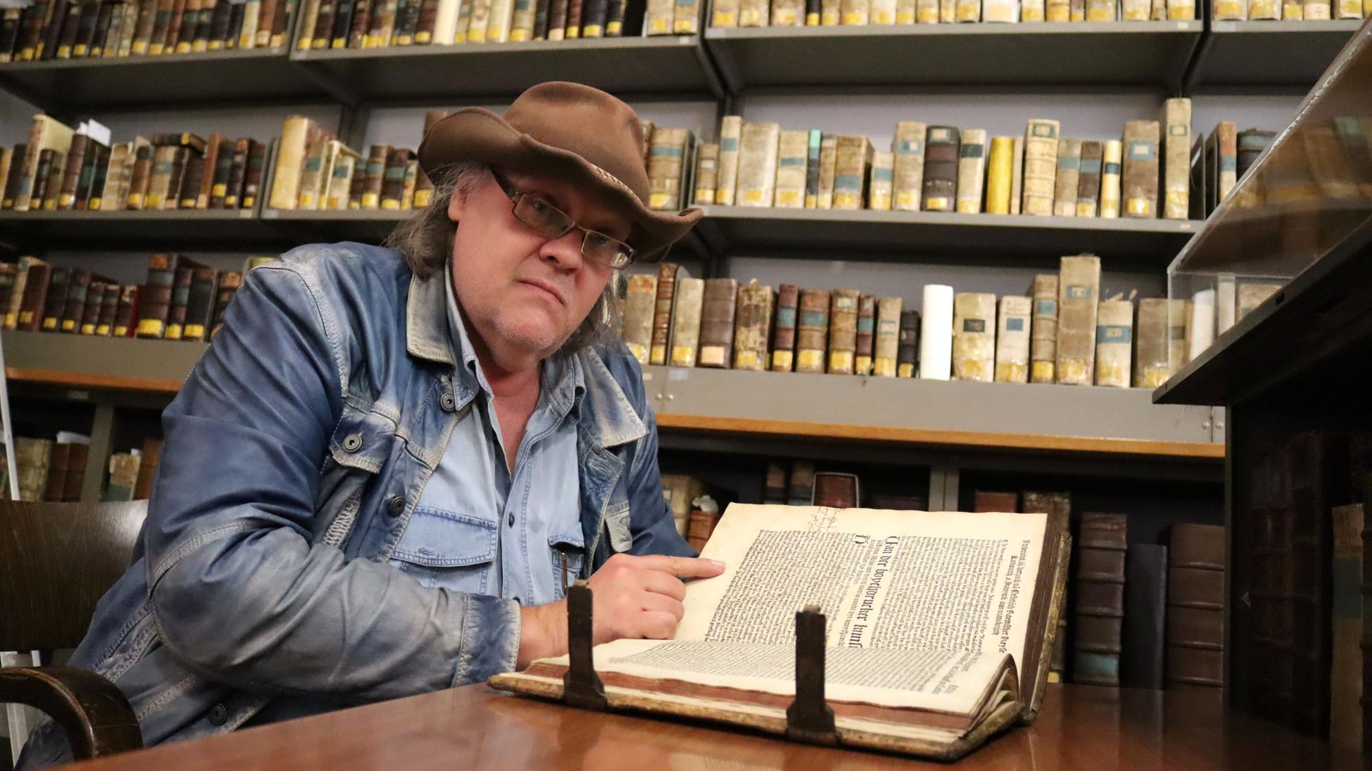 Bongart, der einen Hut trägt, sitzt an einem Tisch, vor sich ein Buch. Im Hintergrnd Regale.