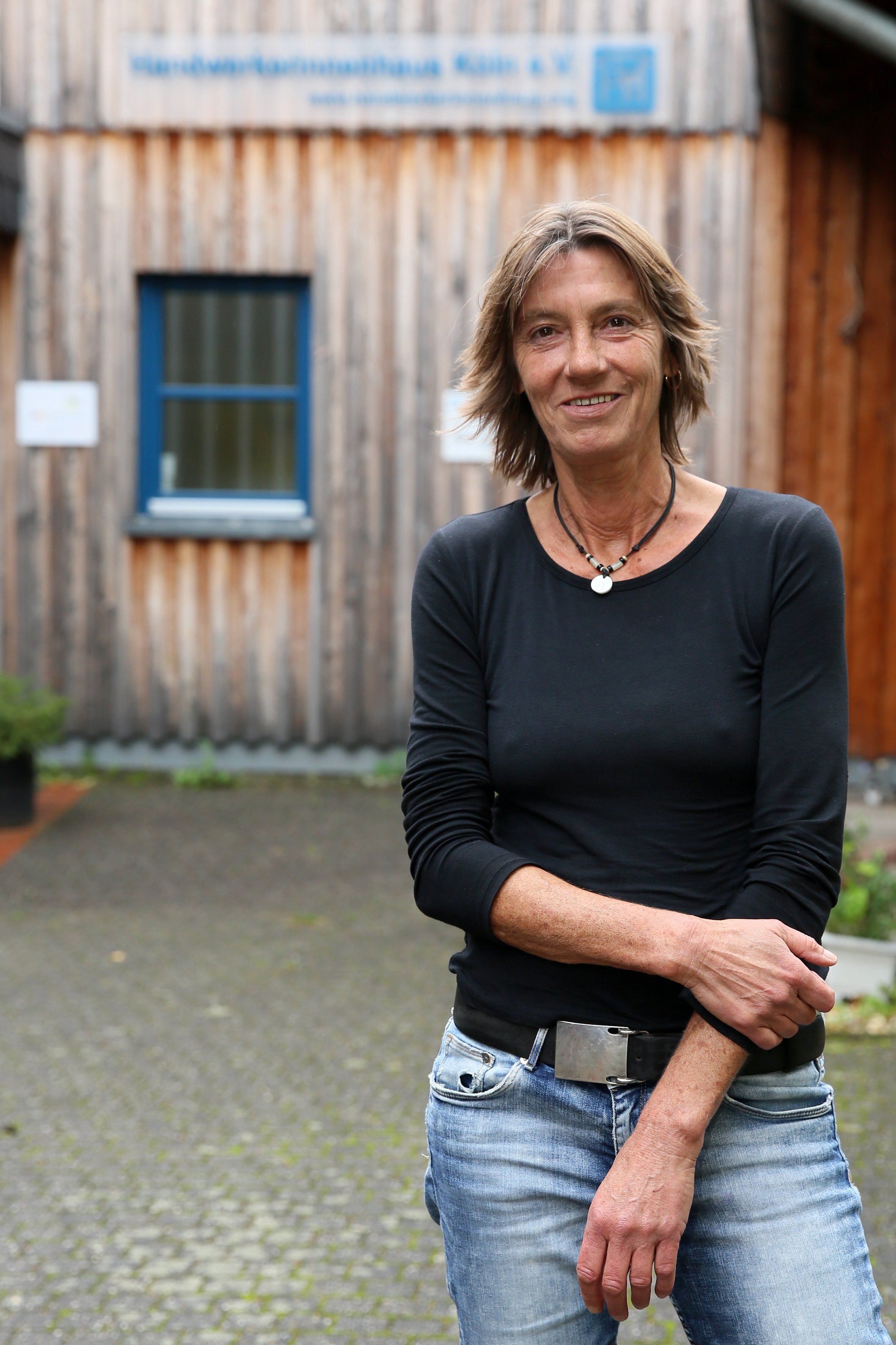 Eine Frau in Jeans und schwarzem Pullover steht vor einem Gebäude aus Holz.