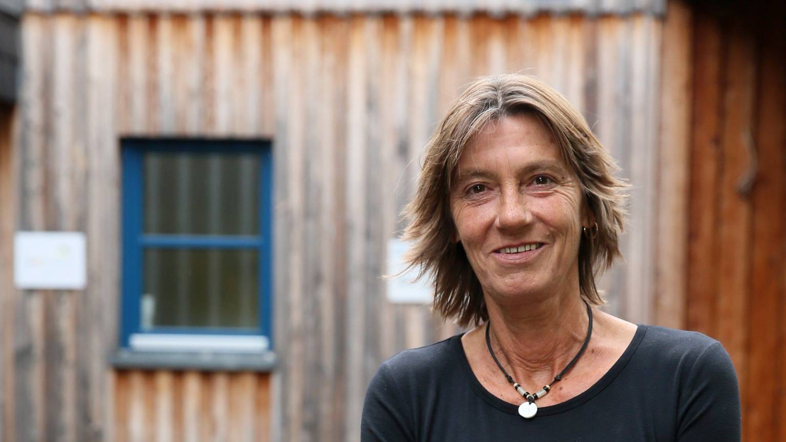 Eine Frau in Jeans und schwarzem Pullover steht vor einem Gebäude aus Holz.