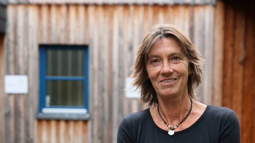 Eine Frau in Jeans und schwarzem Pullover steht vor einem Gebäude aus Holz.