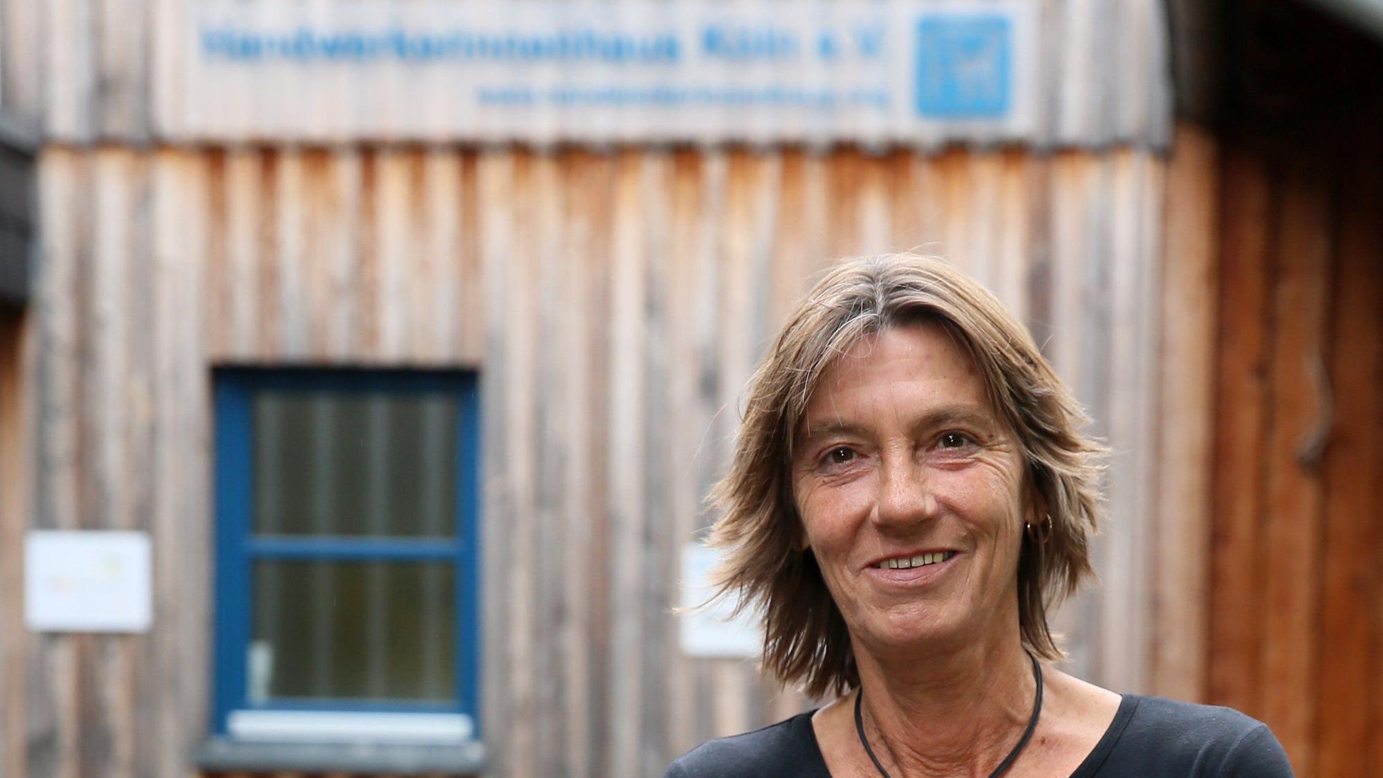Eine Frau in Jeans und schwarzem Pullover steht vor einem Gebäude aus Holz.