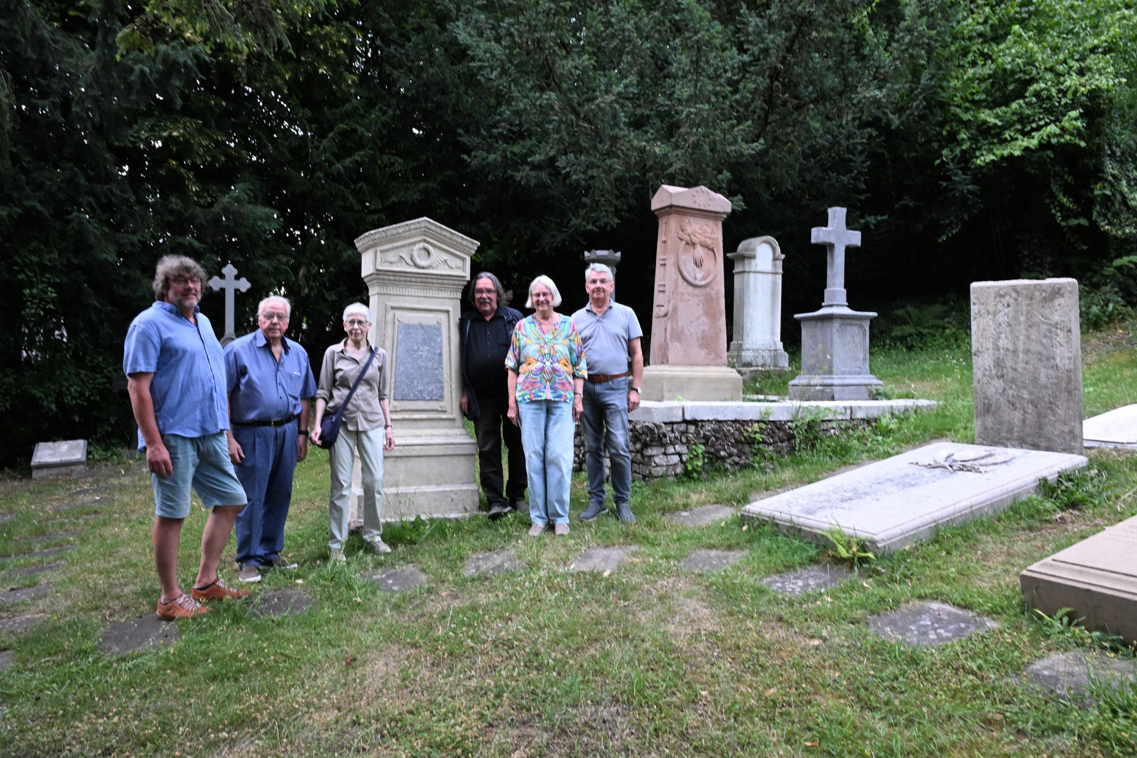 Vier Männer und zwei Frauen stehen auf einem Friedhof vor uralten Grabsteinen udn Grabplatten. Dazwischen Wiese.