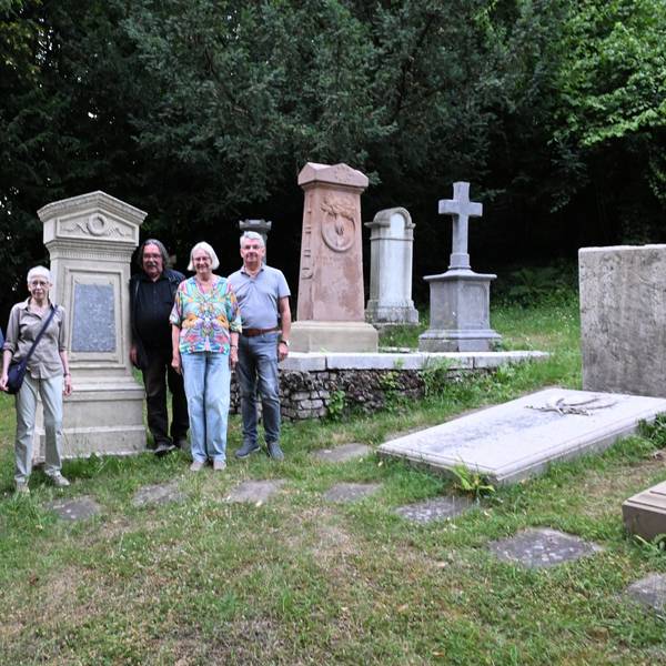 Vier Männer und zwei Frauen stehen auf einem Friedhof vor uralten Grabsteinen udn Grabplatten. Dazwischen Wiese.