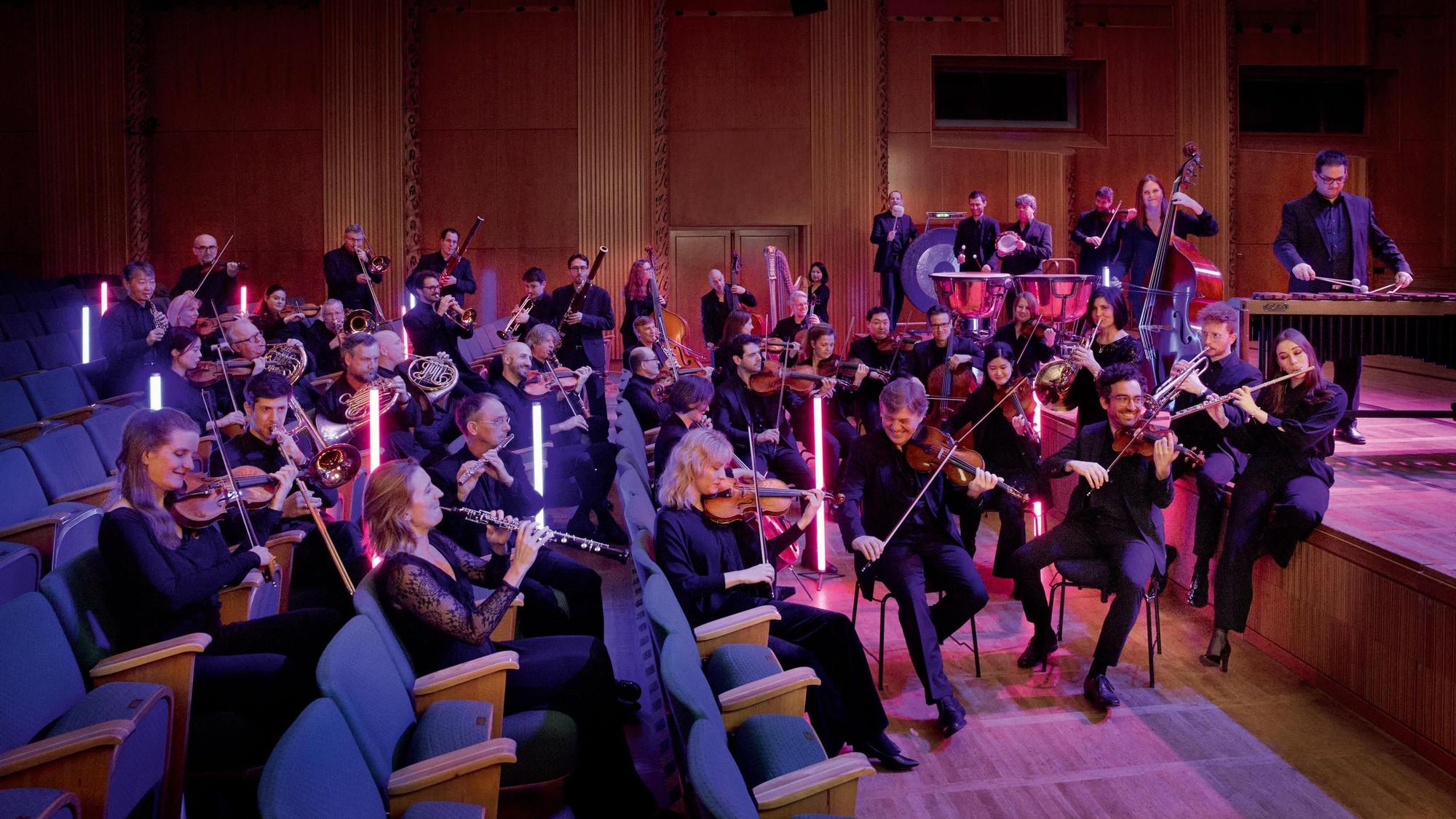 Das Ensemble des WDR-Funkhausorchesters sitzt in den Stuhlreihen eines Konzerthauses.