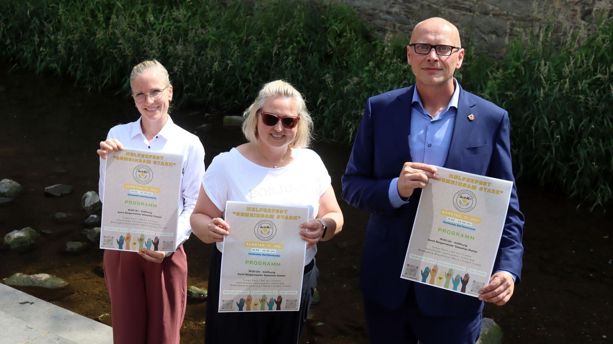 Bürgermeister Sebastian Glatzel (v.r.), Fachbereichsleiterin Stephanie Schmitz und Lisa Eckardt stehen an der Erft und halten Plakate für das Helferfest in den Händen.