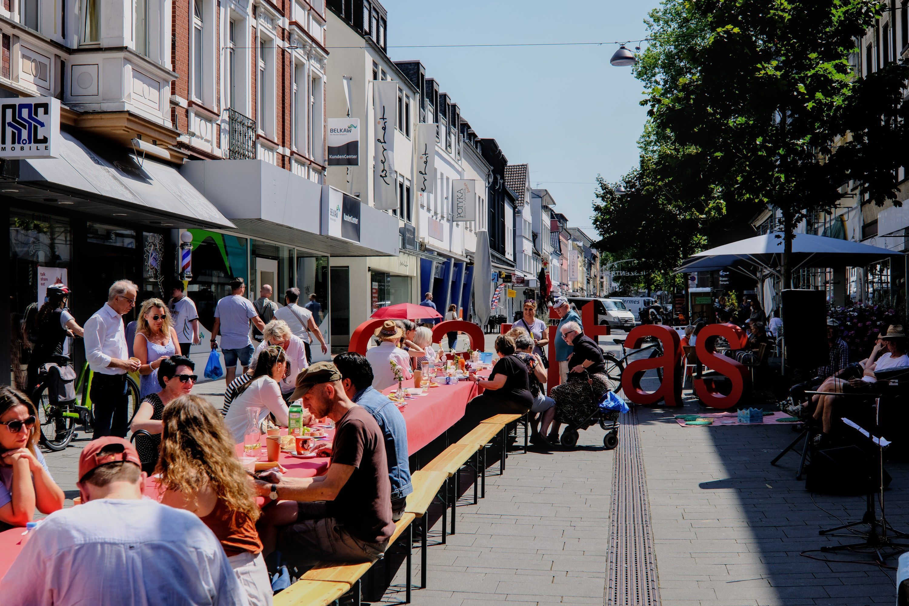 ein mit einer roten Decke gedeckter langer Tisch, daran auf Bierbänken rechts und links viele Menschen im Sonnenschein