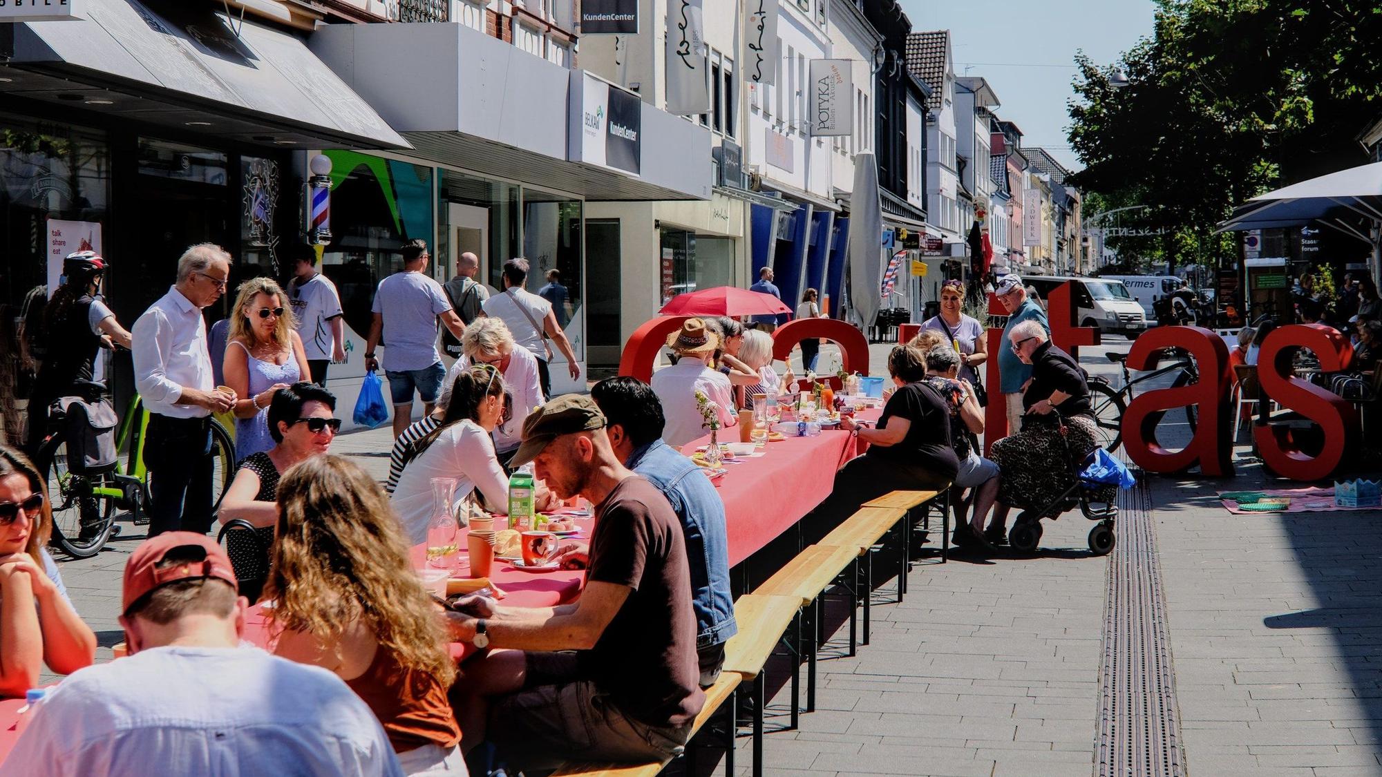 ein mit einer roten Decke gedeckter langer Tisch, daran auf Bierbänken rechts und links viele Menschen im Sonnenschein