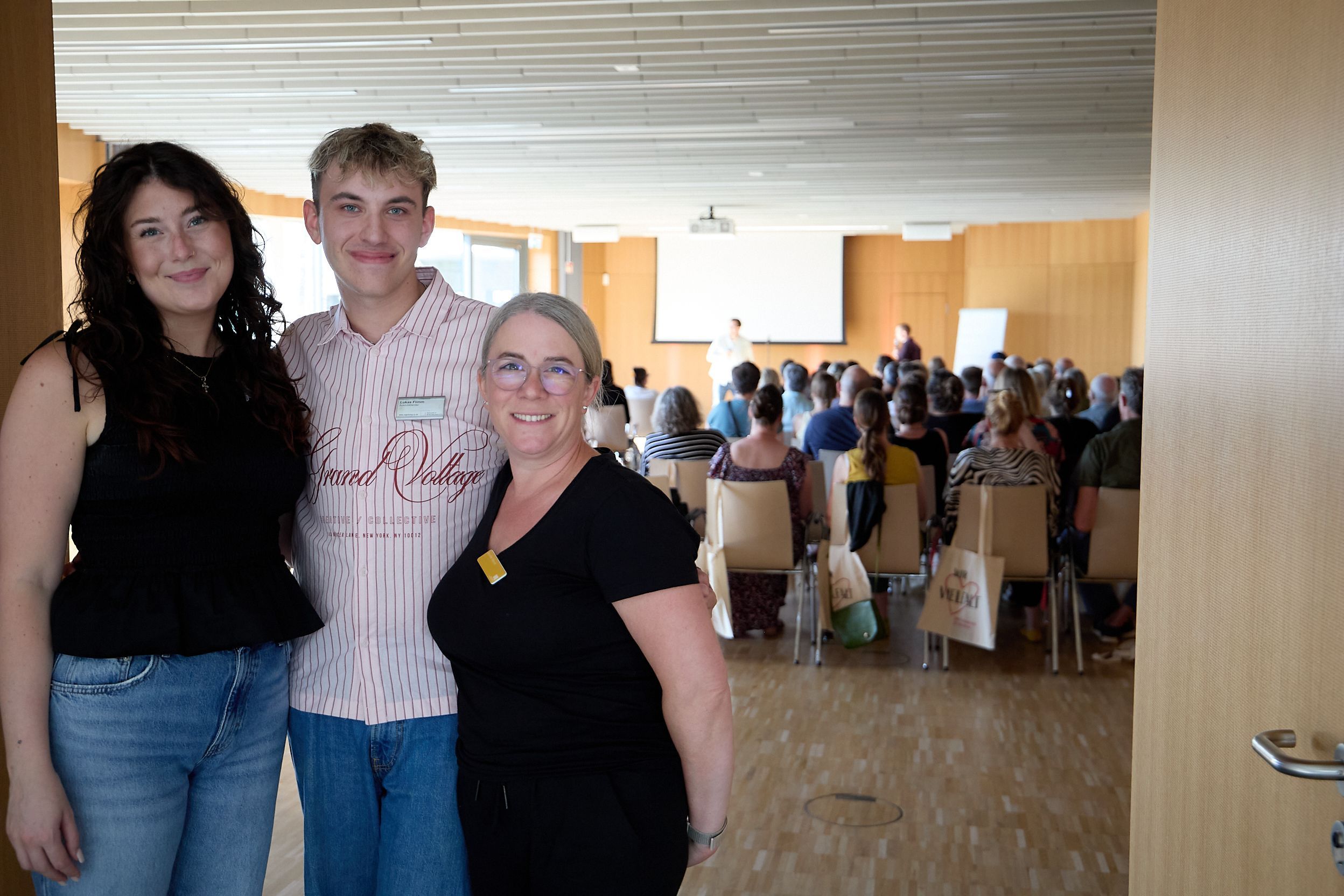 Lukas Flimm, Jennifer Wenck (rechts) und Anna-Lena Stockem stehen vor einem Saal, in dem viele Leute sitzen.