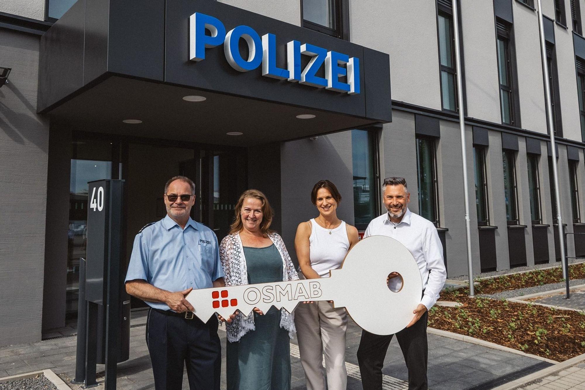 Auf dem Bild sind zwei Frauen und zwei Männer vor dem neuen Polizeigebäude zu sehen, sie halten einen großen symbolischen Schlüssel in der Hand.