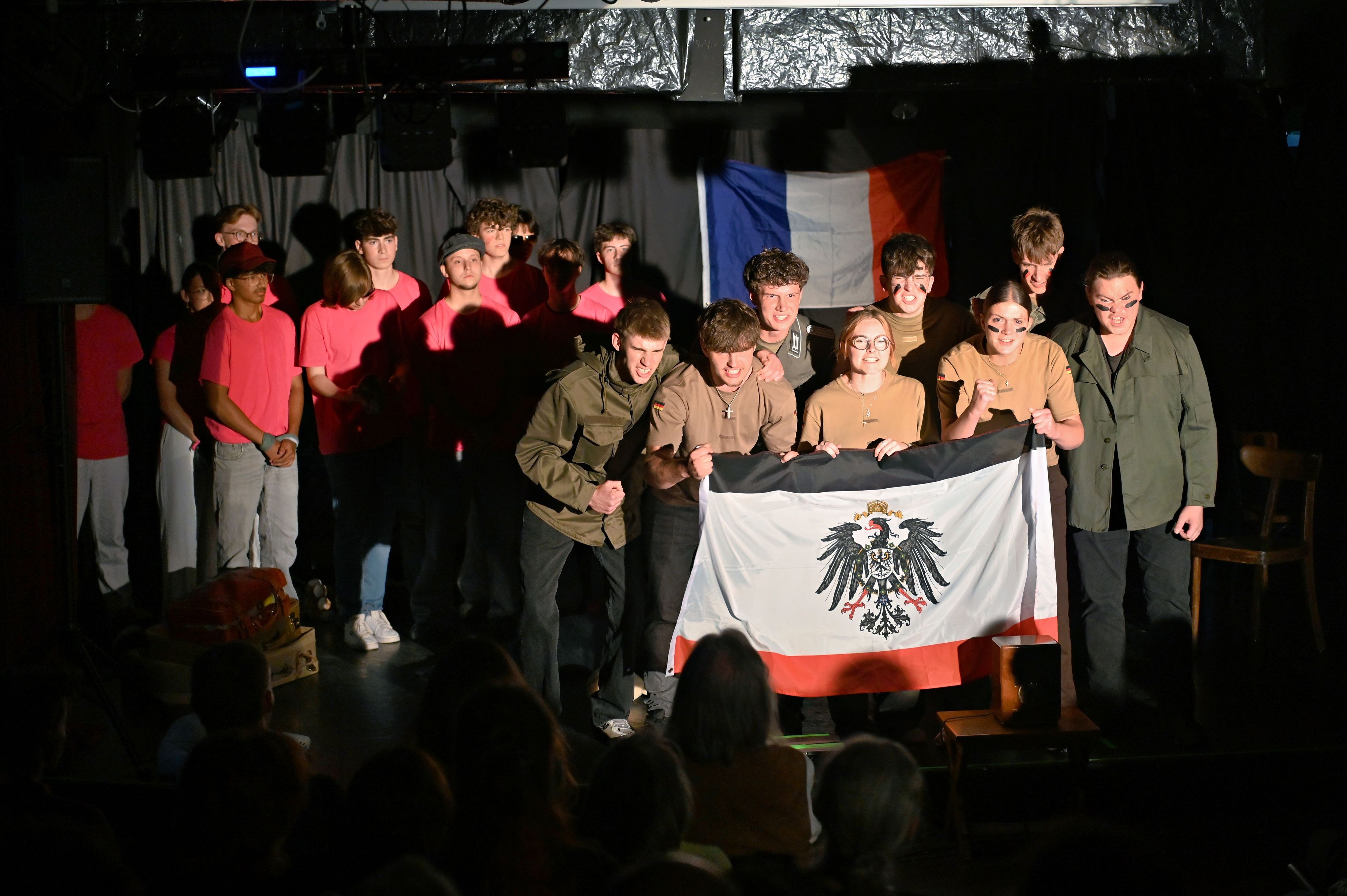 Die Schülerinnen und Schüler bilden auf der Bühne zwei Gruppen. Die einen tragen T-Shirts in Pink, die anderen sind in Oliv gekleidet und zeigen eine Flagge mit dem Reichsadler.