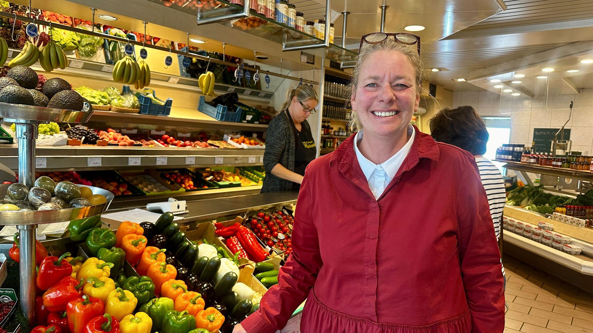 Eine Frau in roter Jacke mit der hochgeschobenen Brille im Haar steht neben einem Aufsteller mit Obst und Gemüse.