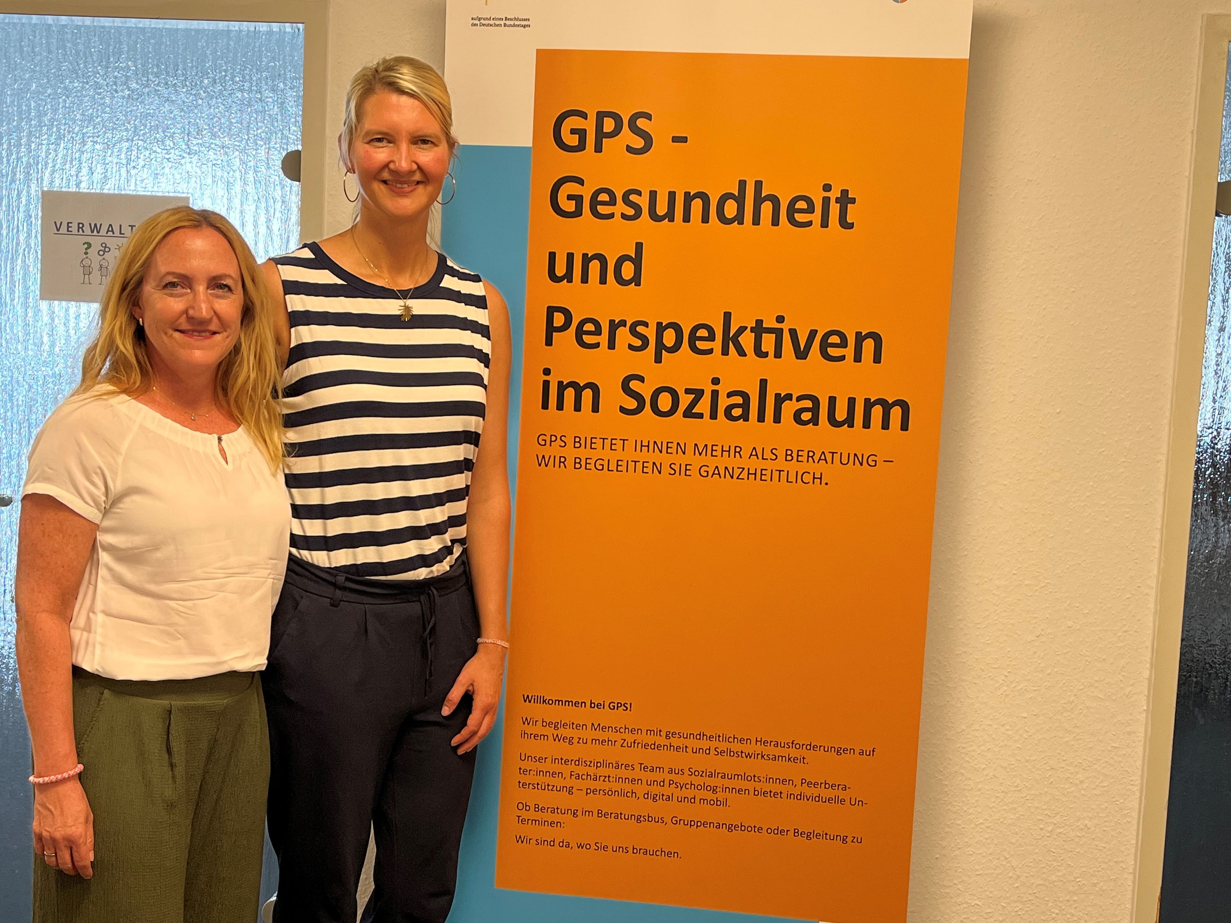 Die beiden Frauen stehen neben einer Werbetafel.