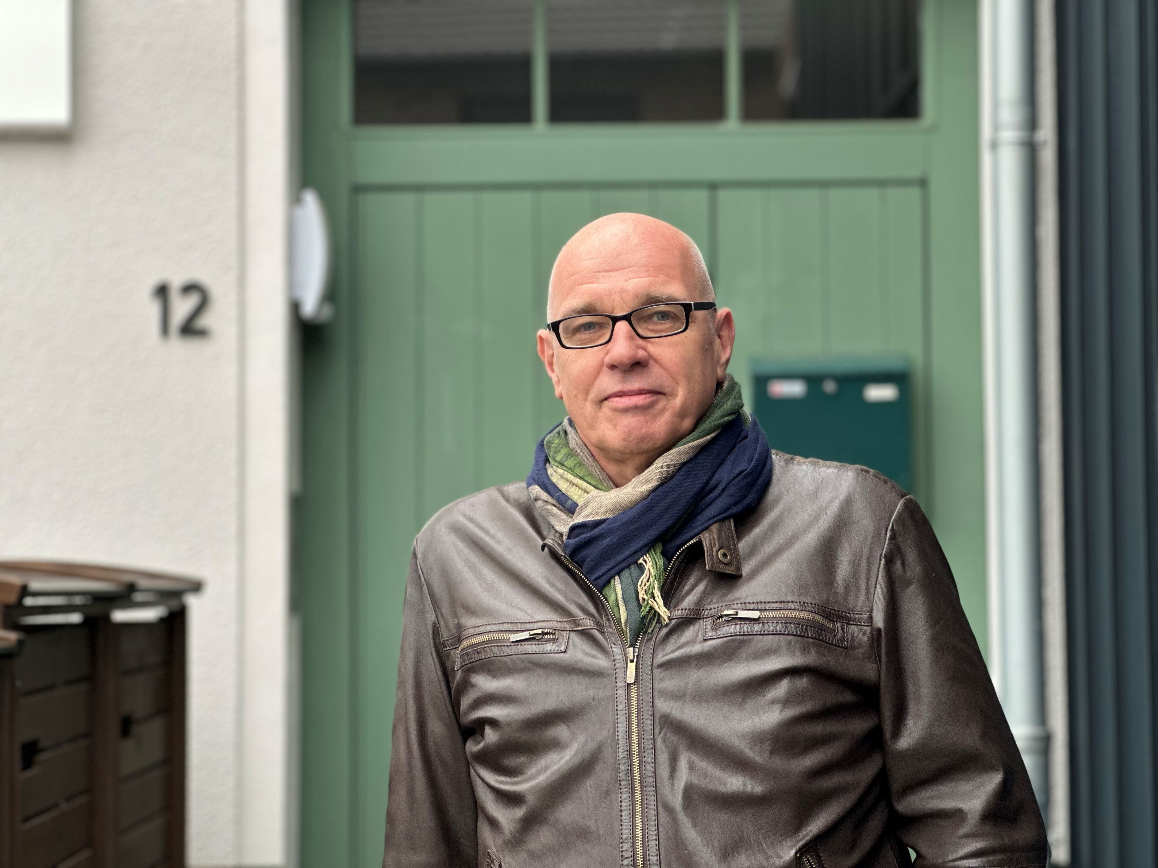 Ein Mann mit Glatze und Brille steht vor einem Haus mit grüner Tür.