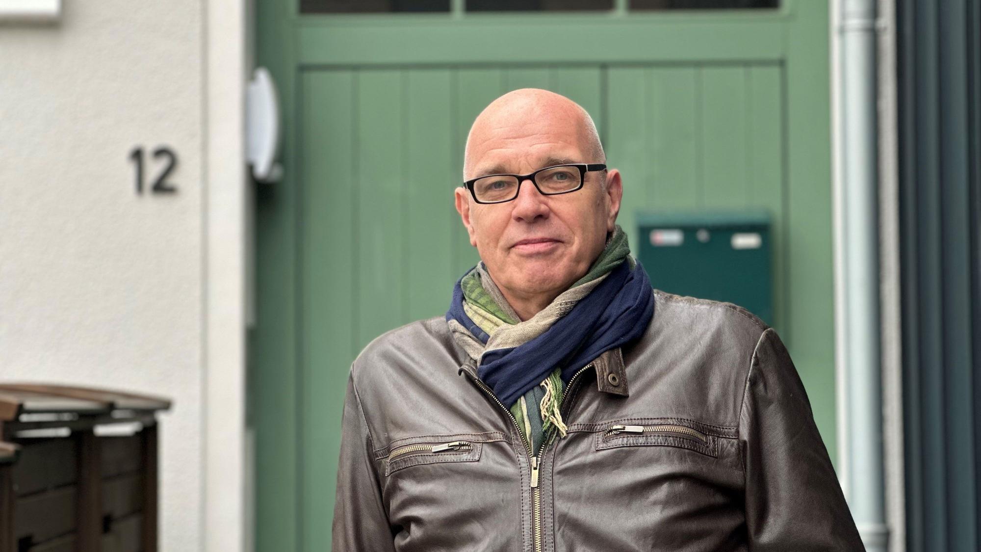 Ein Mann mit Glatze und Brille steht vor einem Haus mit grüner Tür.