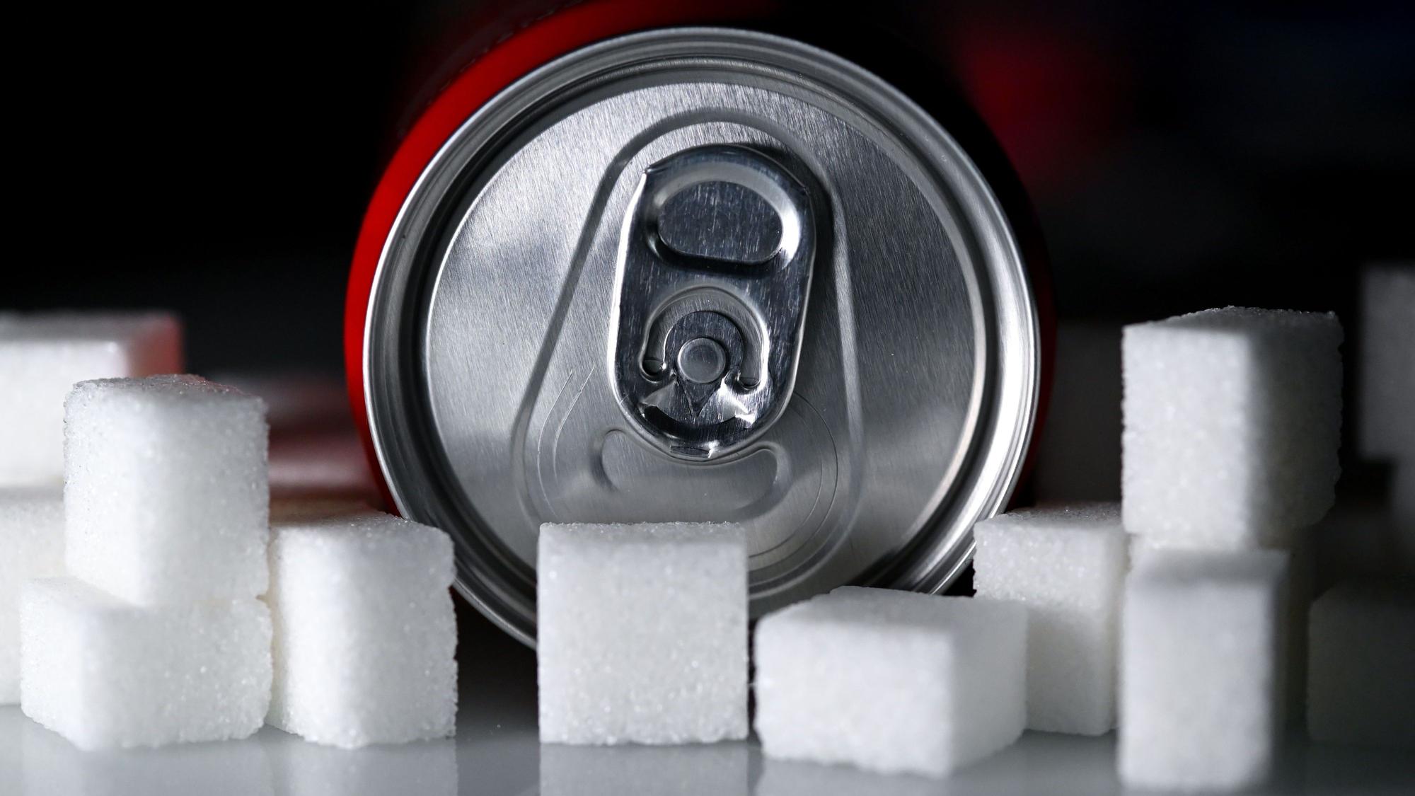 Eine Getränkedose mit einem Softdrink und zahlreiche Zuckerwürfel liegen auf einem Tisch. Die Bundesregierung plant eine Zuckersteuer. (Symbolbild)