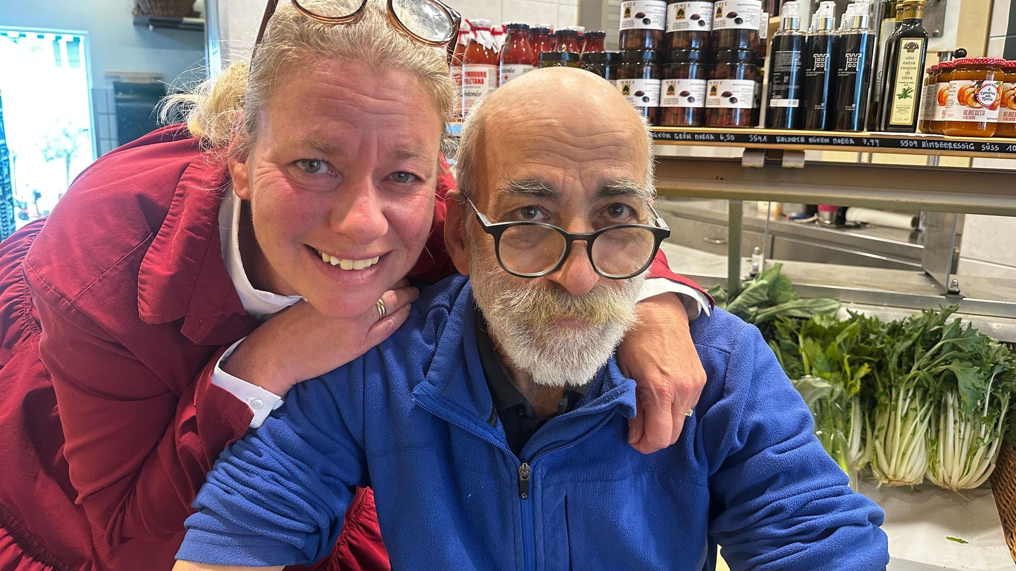 Eine Frau mit roter Jacke und Brille im Haar umarmt einen älteren Mann mit Bart, blauer Jacke und Brille.