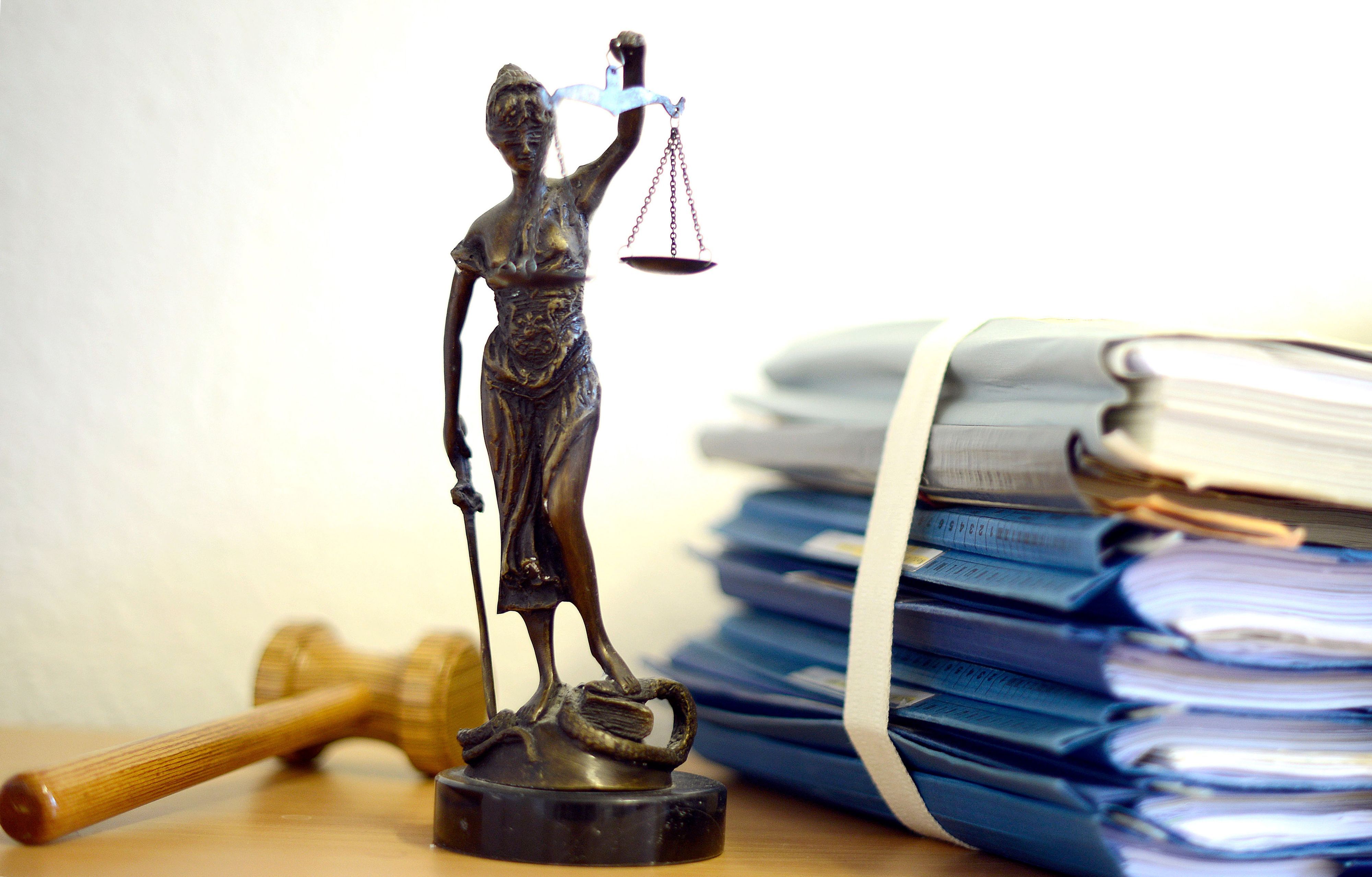 Die modellhafte Nachbildung der Justitia steht in einem Raum eines Richters neben einem Holzhammer und einem Aktenstapel.