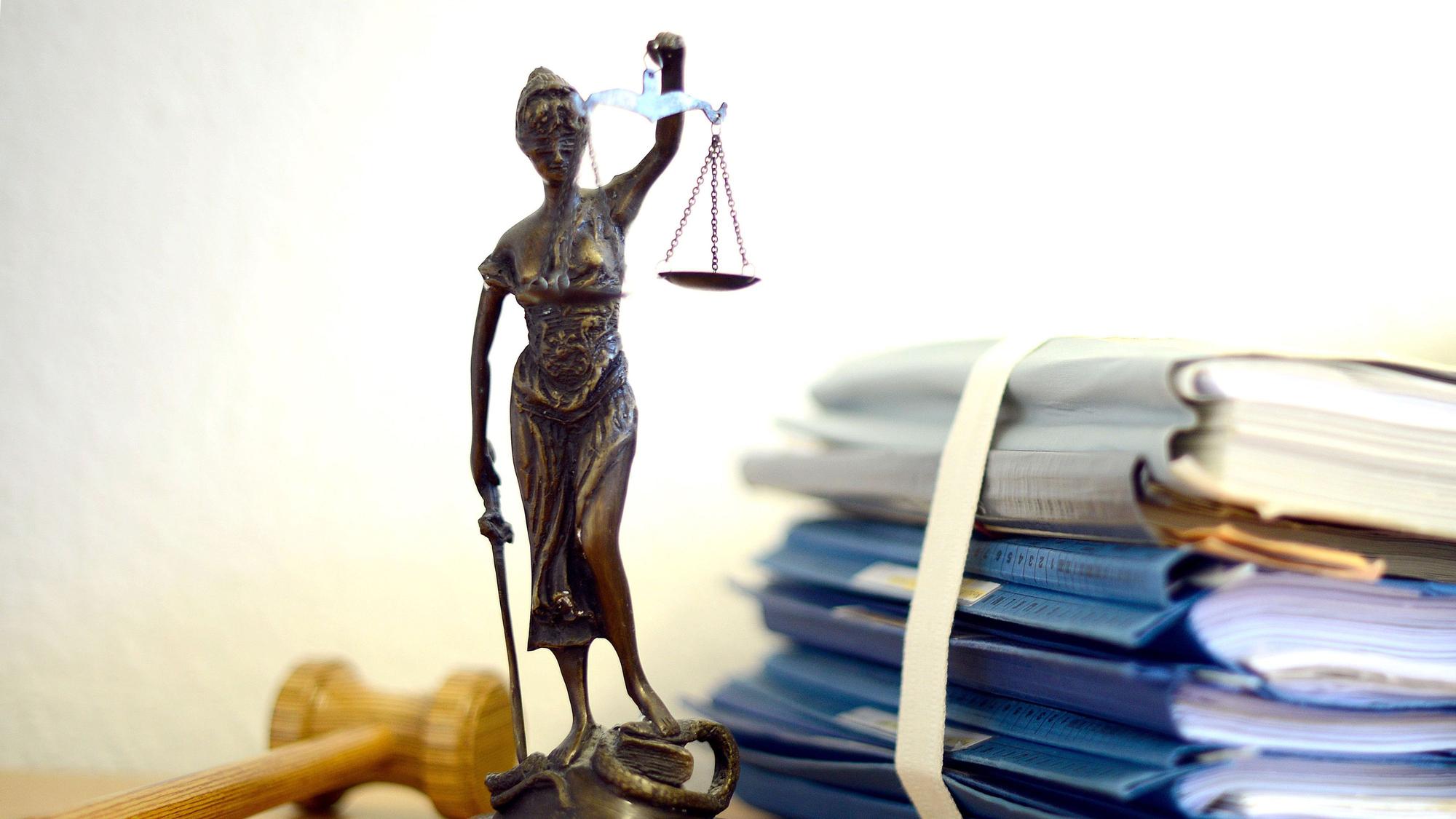 Die modellhafte Nachbildung der Justitia steht in einem Raum eines Richters neben einem Holzhammer und einem Aktenstapel.