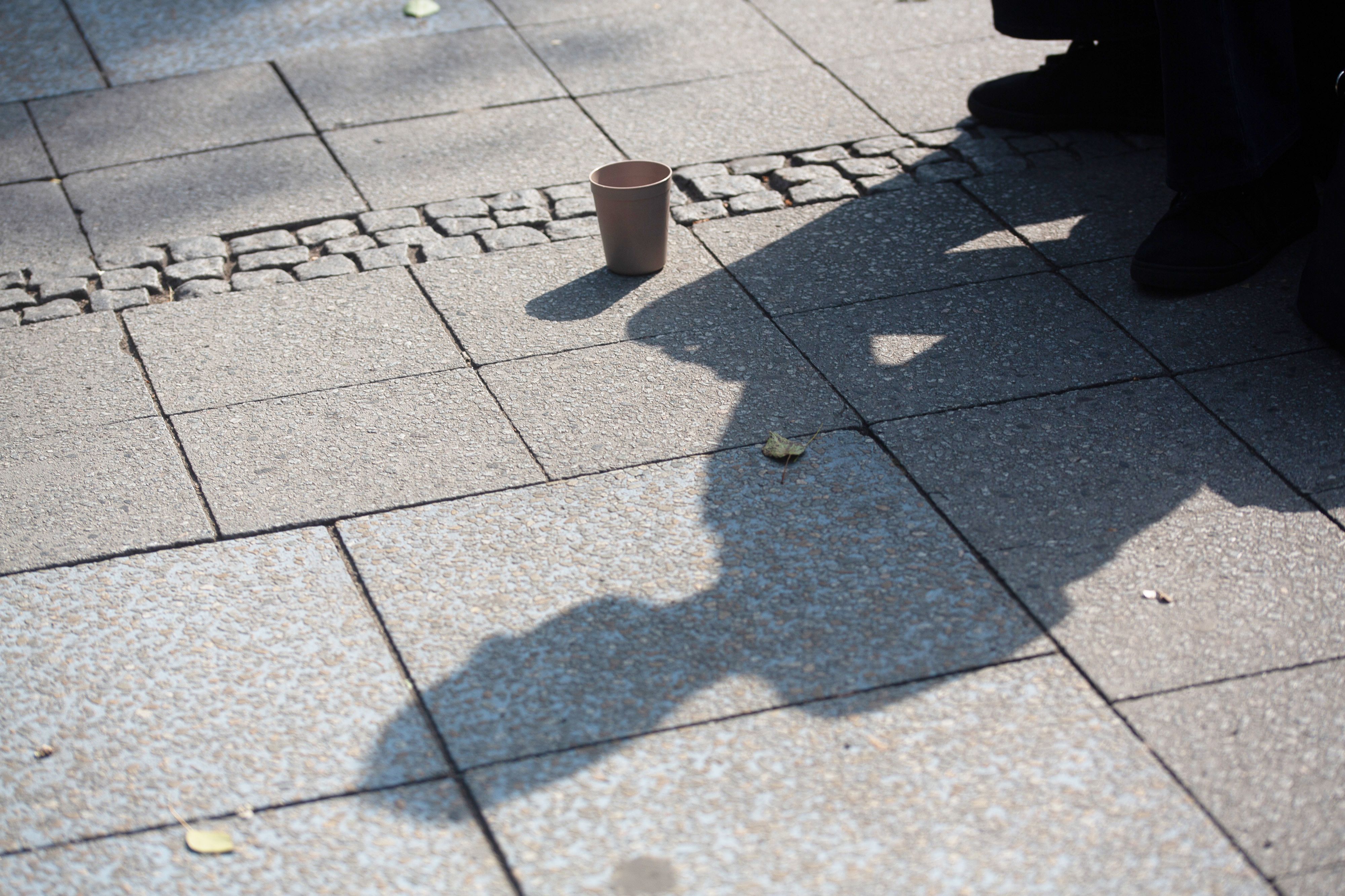 SYMBOLBILD - 10.07.2023, Berlin: Der Schatten einer Person mit einem Becher für Spenden fällt auf den Gehweg. (zu dpa: «Wohnungslosenhilfe fordert Trinkwasserstellen für Obdachlose») Foto: Fernando Gutierrez-Juarez/dpa +++ dpa-Bildfunk +++