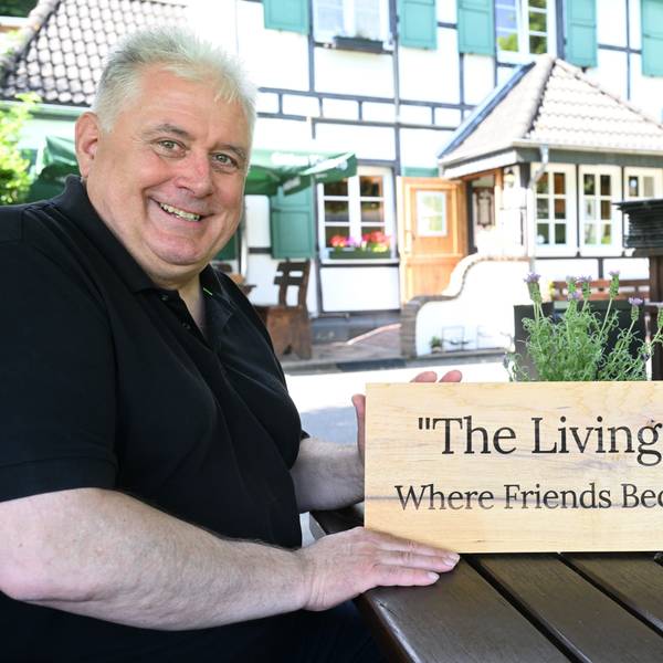 Ein Mann sitzt vor einem bergischen Fachwerklokal und hält ein Holzschild mit der Aufschrift: „The Living Room. Where Friends Become Family“ in der Hand.