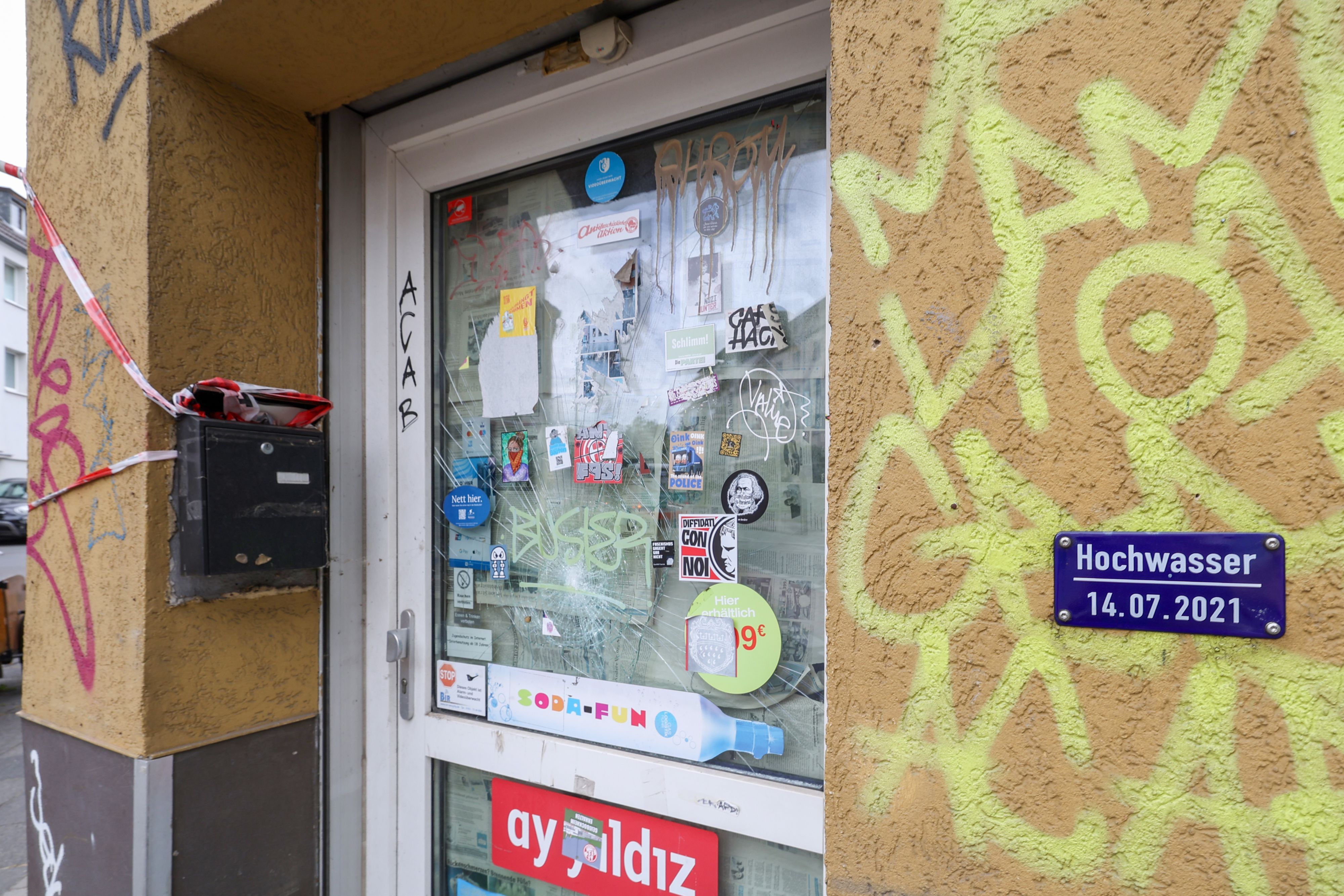 Eine Tür zu einem Kiosk ist zu sehen. Das Haus ist besprüht mit Graffiti. Eine Hochwassermarke ist an der Hauswand angebracht.