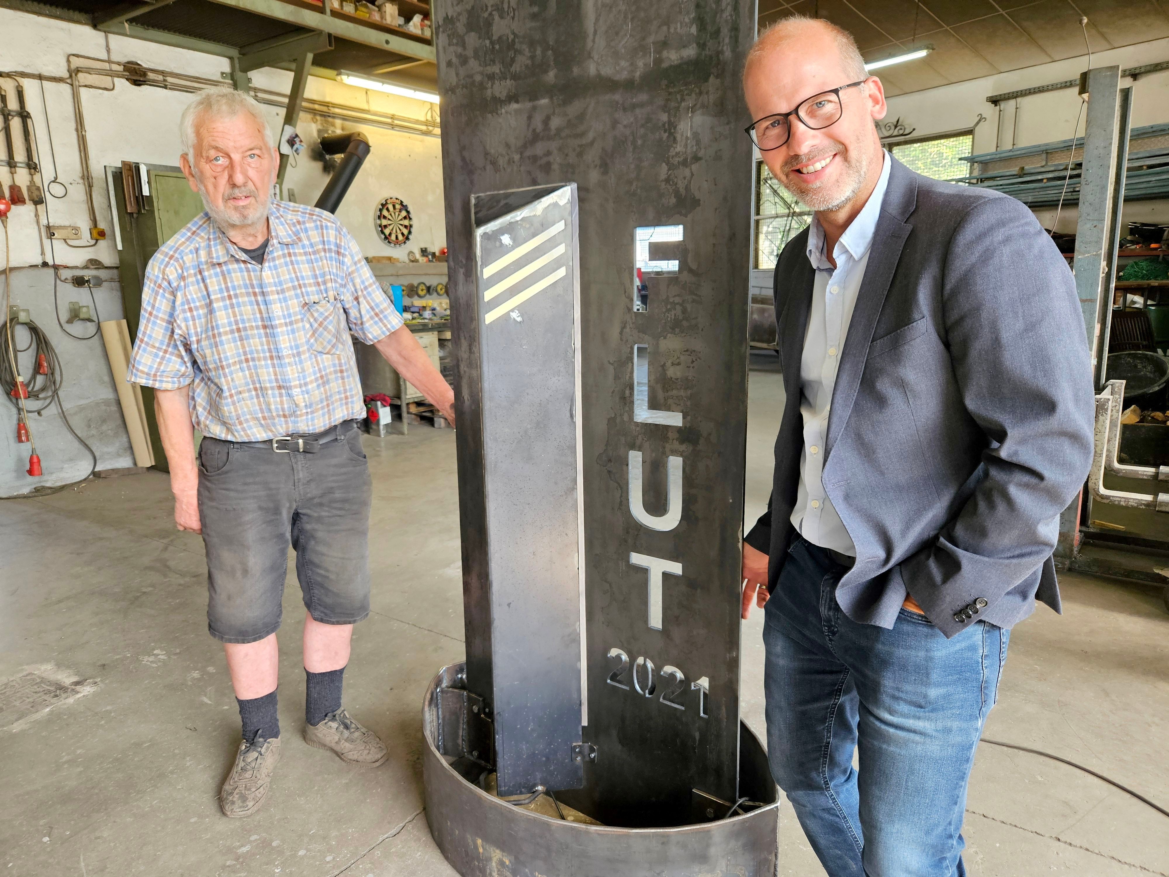 Zwei Männer stehen neben einer Metallstele, in die „Flut 2021“ eingraviert ist.