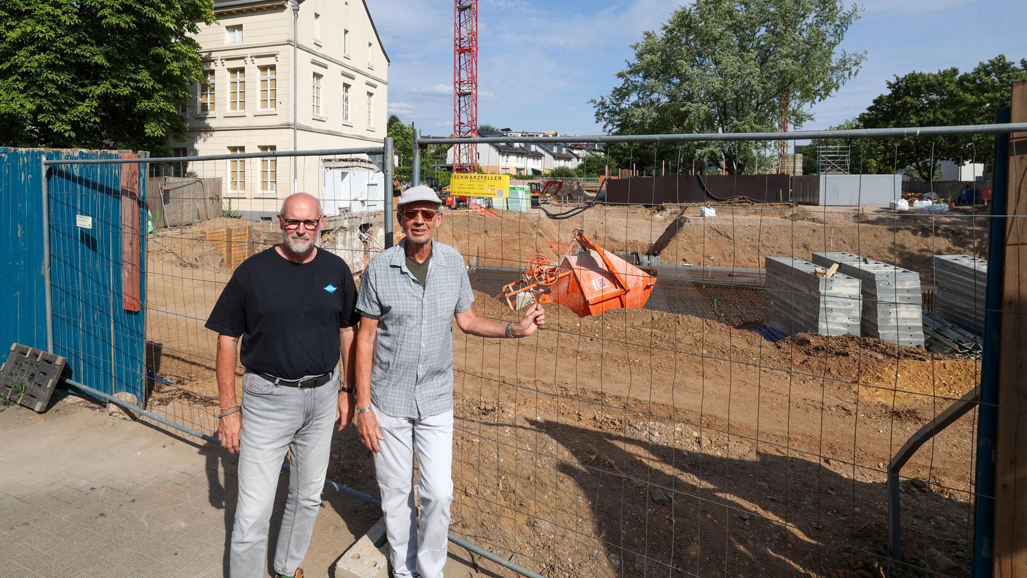 Zwei Männer stehen nebeneinander vor einem Bauzaun. Dahinter sind die Baustelle, ein Kran, und ein Gutshaus zu sehen.