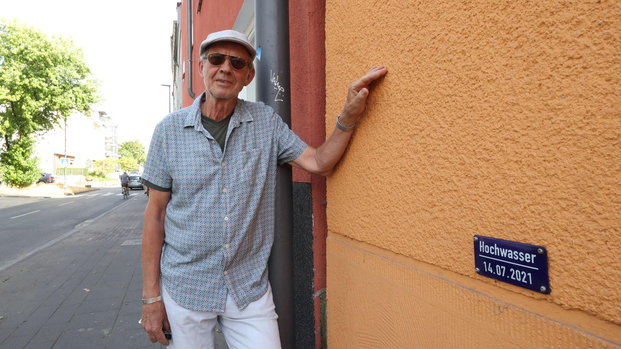 Ein Mann mit Kappe und Sonnenbrille steht vor einer Hauswand. Auf dieser ist eine Hochwassermarke angebracht.
