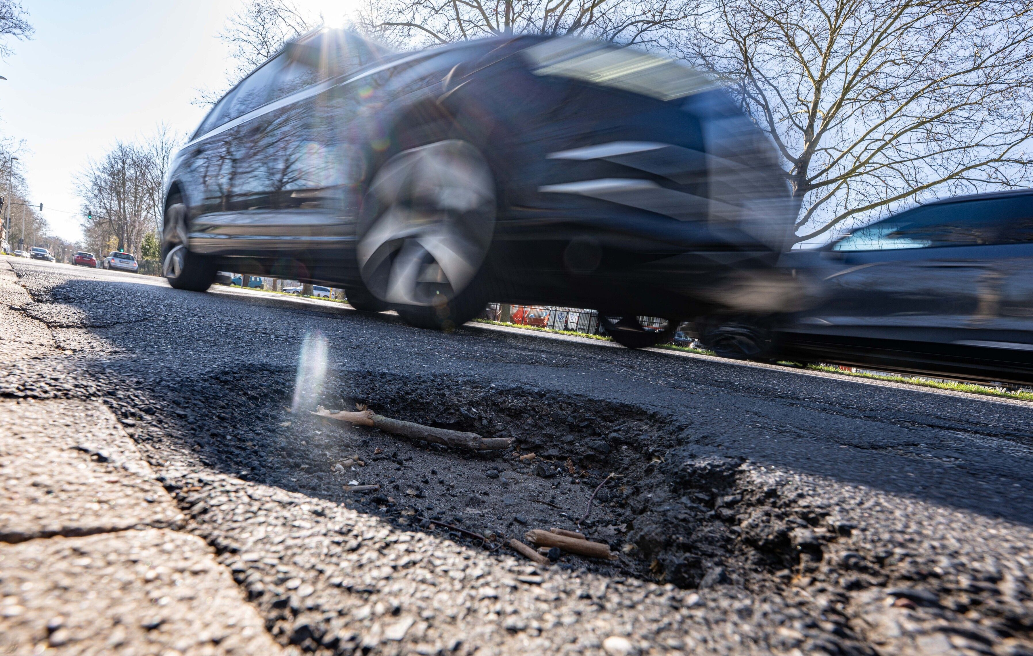 Das Foto zeigt ein Schlagloch, dahinter fährt ein Auto über die Straße.