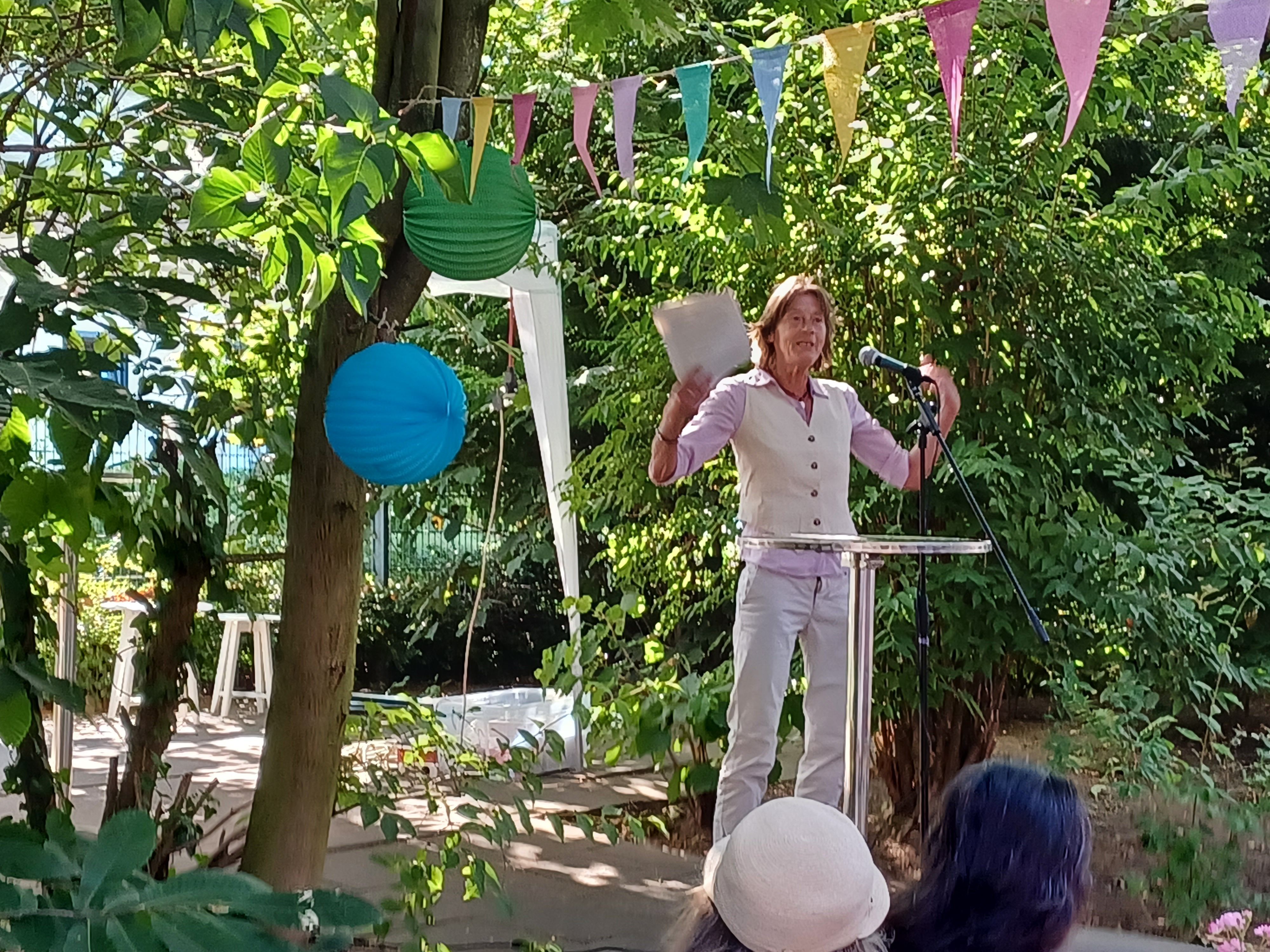 Eine Frau in rosa Hemdbluse mit Weste darüber steht an einem Stehtisch draußen in einem Garten und spricht in ein Mikrofon.