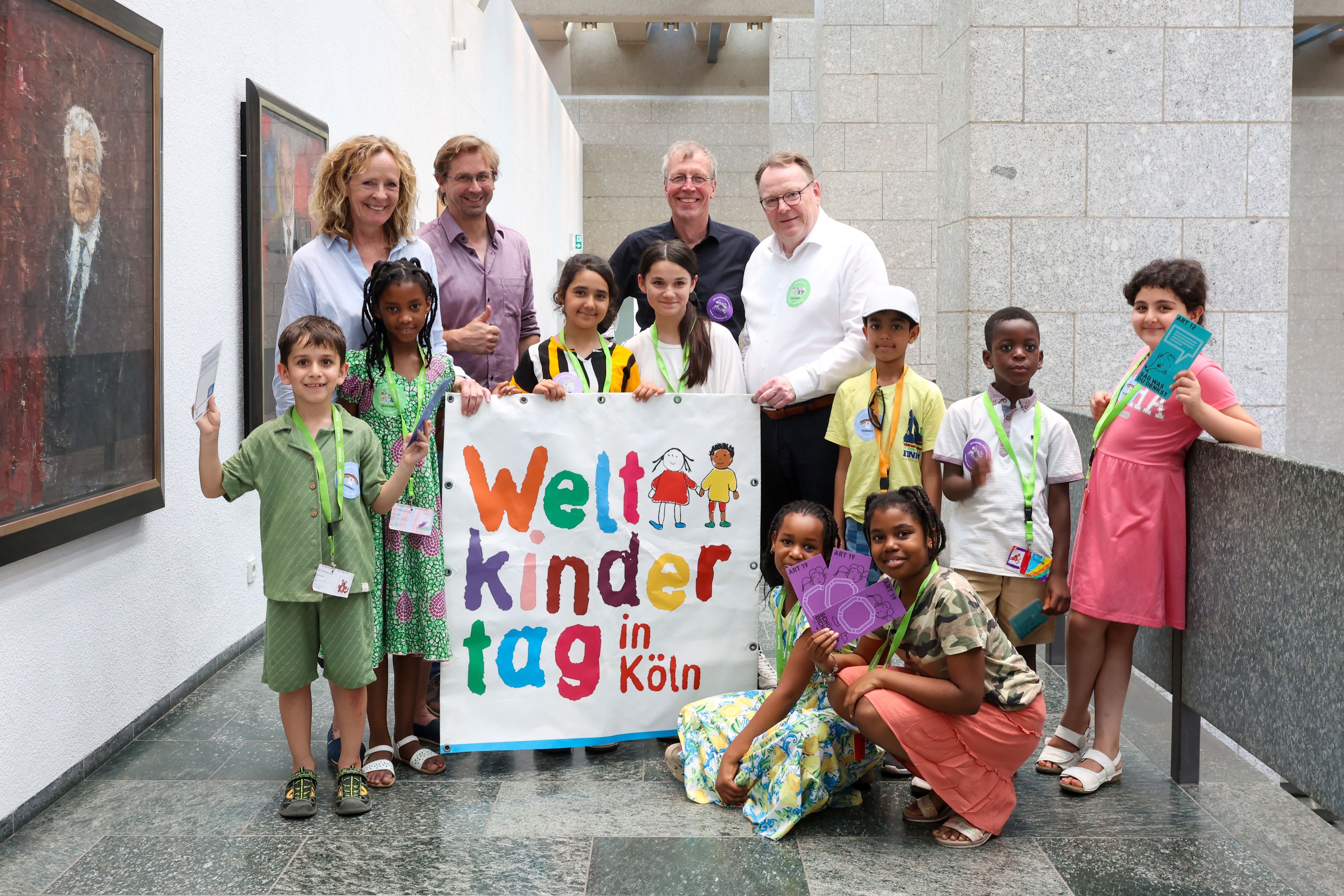 Neun Jungen und Mädchen des Linoclub-Kinderrates stehen gemeinsam mit Isabella Neven DuMont (wir helfen e.V.), Lars Hüttler (Kinderschutzbund Köln), Hans-Josef Saxler (Soziales Zentrum Linoclub) und Oberbürgermeister Torsten Burmester im Flur des Historischen Rathauses. Sie halten einen großes Weltkindertag-Plakat in den Händen.