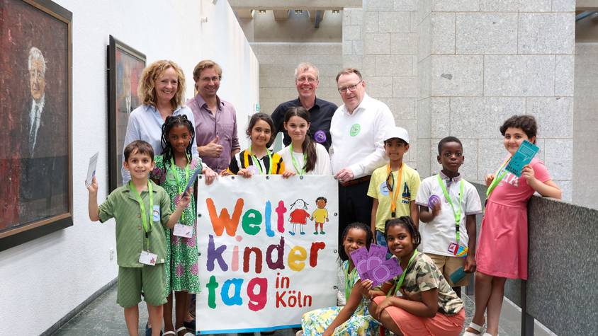 Neun Jungen und Mädchen des Linoclub-Kinderrates stehen gemeinsam mit Isabella Neven DuMont (wir helfen e.V.), Lars Hüttler (Kinderschutzbund Köln), Hans-Josef Saxler (Soziales Zentrum Linoclub) und Oberbürgermeister Torsten Burmester im Flur des Historischen Rathauses. Sie halten einen großes Weltkindertag-Plakat in den Händen.