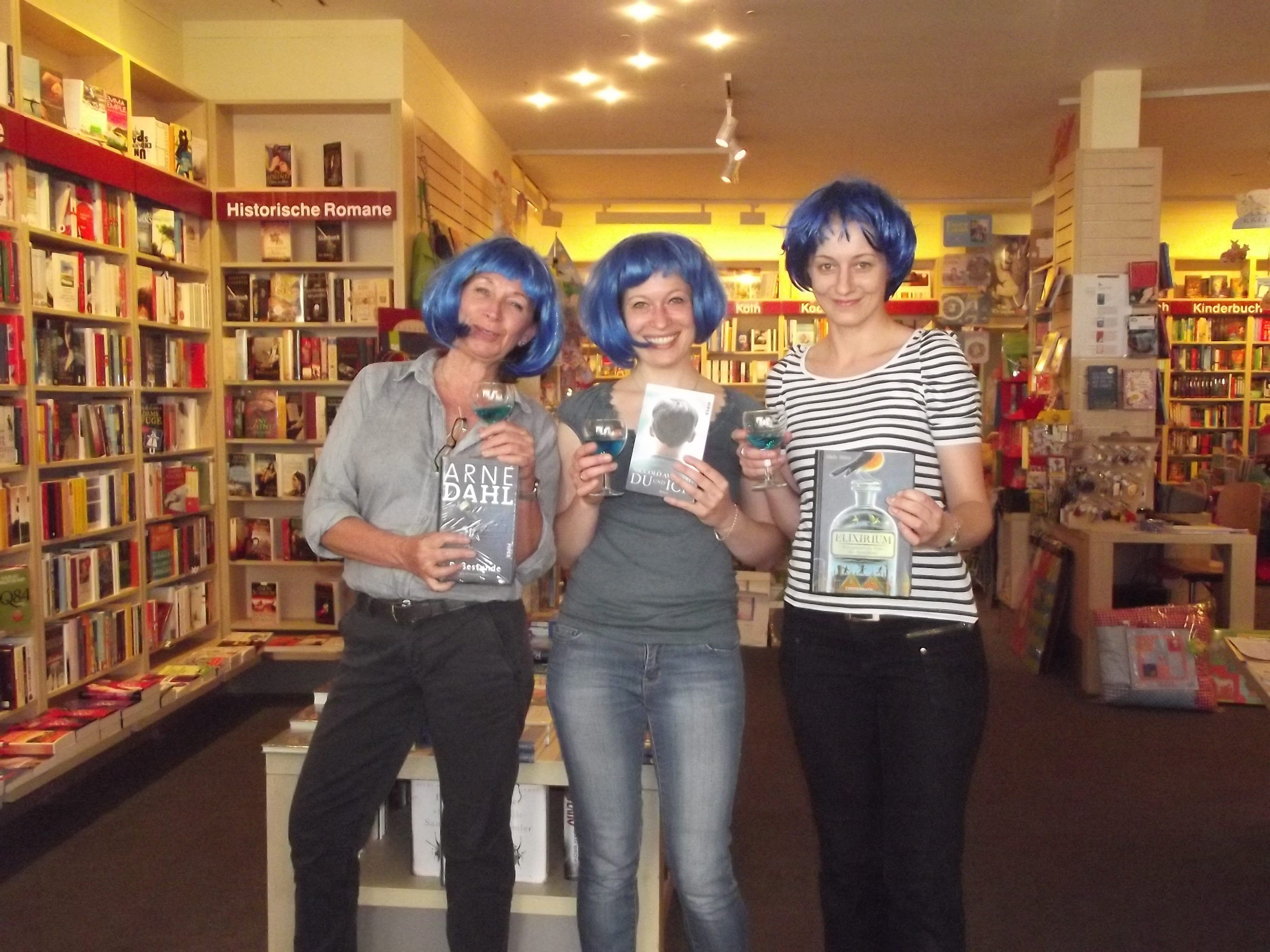 Drei Frauen mit blauen Perücken stehen in einer Buchhandlung nebeneinander.