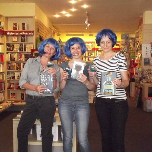 Drei Frauen mit blauen Perücken stehen in einer Buchhandlung nebeneinander.