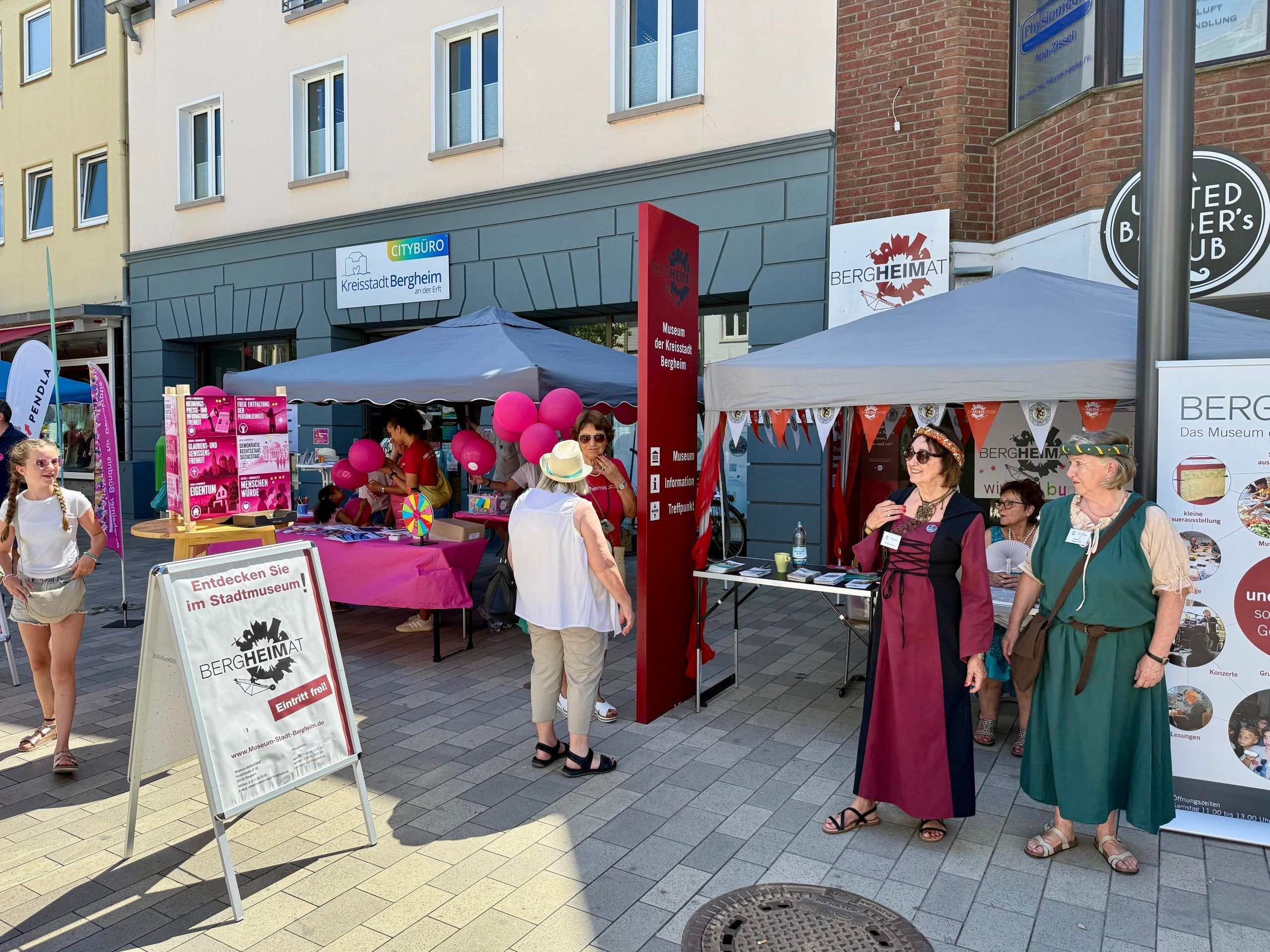 Zu sehen sind teils kostümierte Menschen an einem Stand in einer Fußgängerzone.