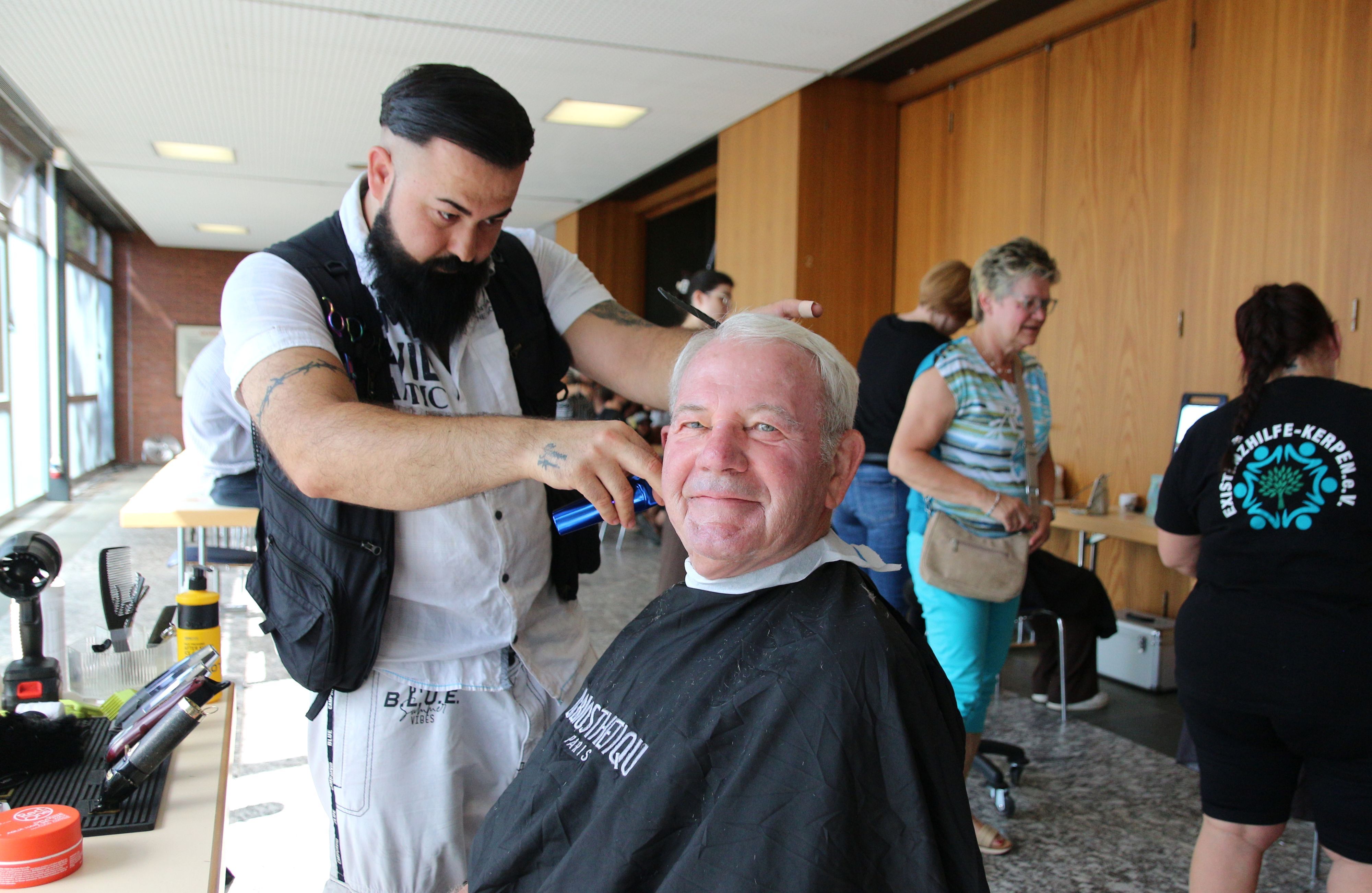 Das Bild zeigt den lächelnden stellvertretender Bürgermeister von Kerpen und den Friseur, der ihm die Haare schneidet. Im Hintergrund weitere Menschen.