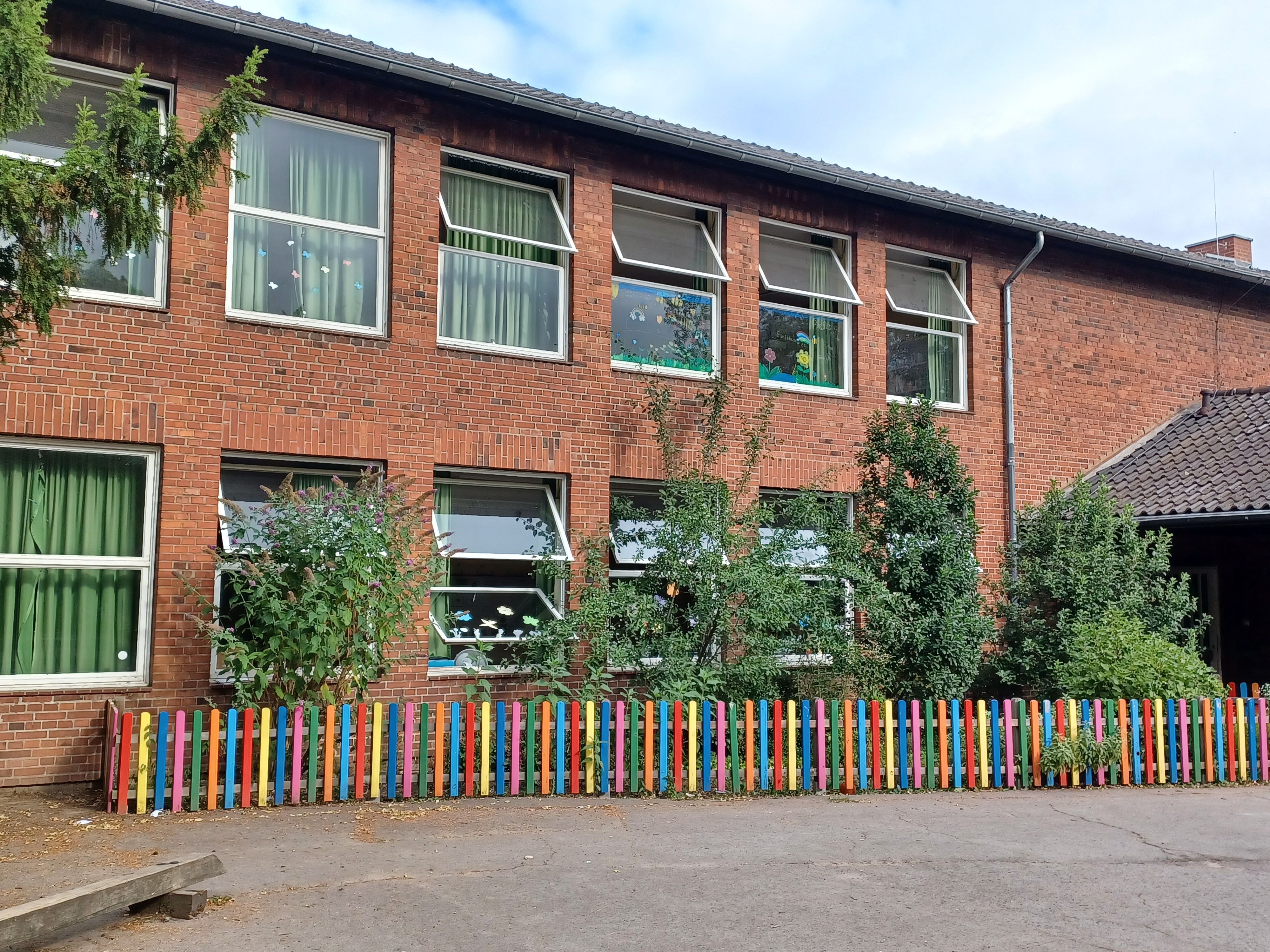 Hinter einem bunten Zaun liegt ein zweigeschossiges Schulgebäude mit großen Fenstern.