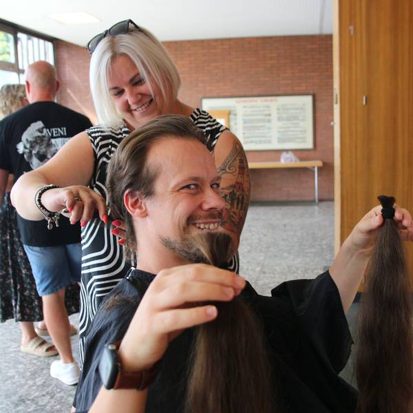 Das Bild zeigt, wie ein Mann seinen abgeschnittene Zöpfe hält und lächelt. Hinter ihm ist die Frisuerin noch an seinen Haaren am Werk.