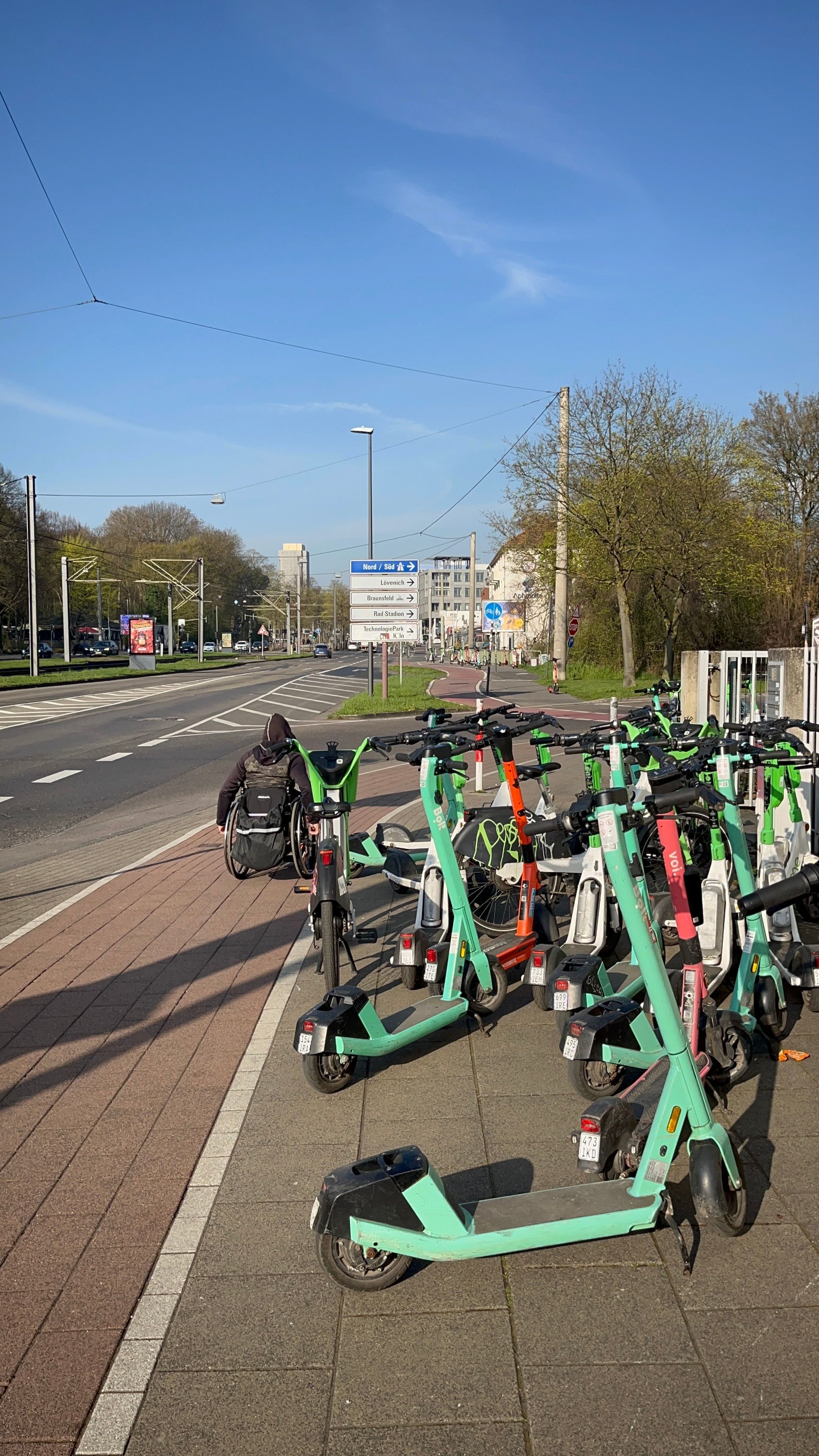 Ein Rollstuhlfahrer muss wegen der auf dem Gehweg abgestellten E-Scooter auf den Radweg ausweichen.
