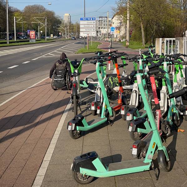 Ein Rollstuhlfahrer muss wegen der auf dem Gehweg abgestellten E-Scooter auf den Radweg ausweichen.